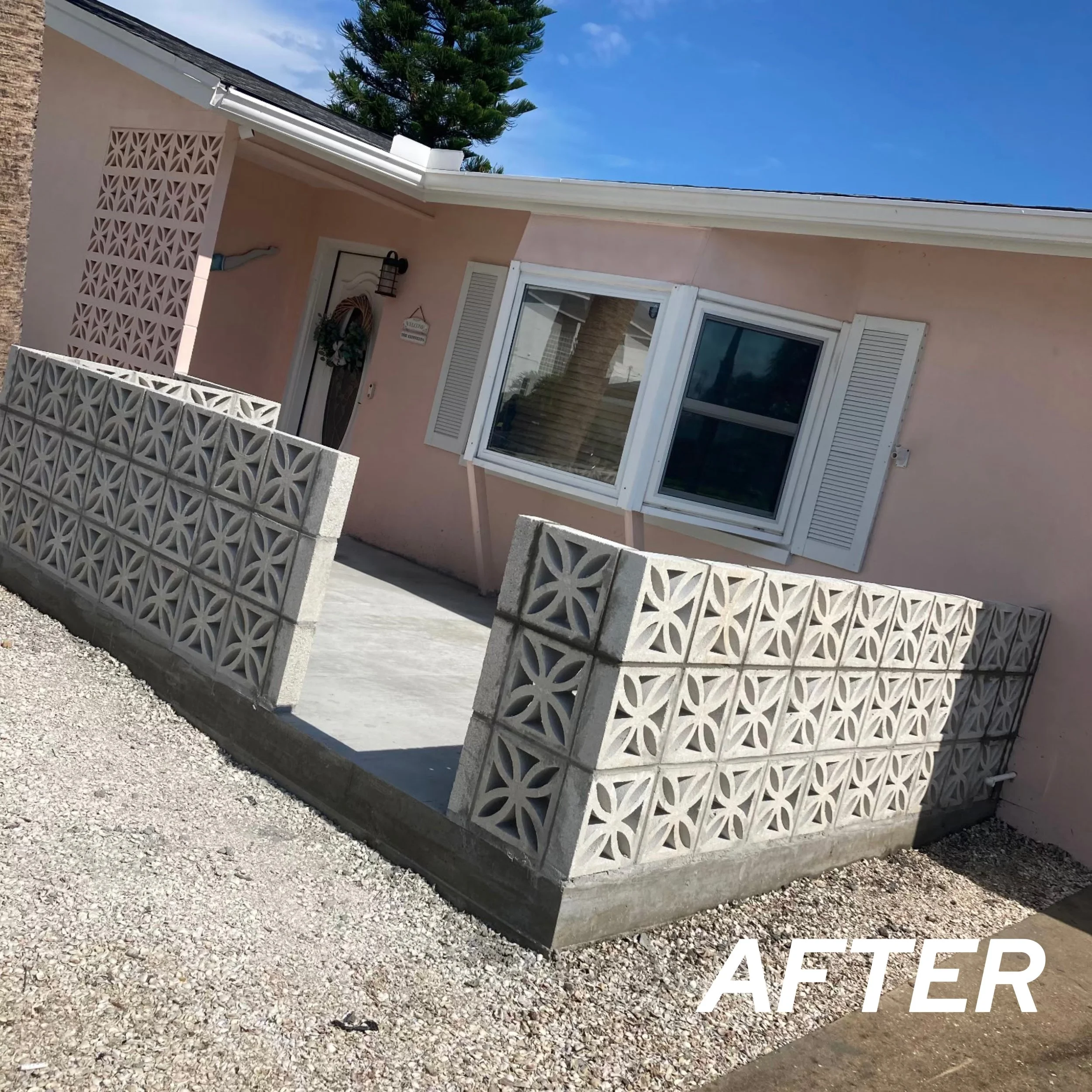 A small pink house with white window shutters and a decorative front door, surrounded by a concrete block fence with geometric patterns, under a blue sky.