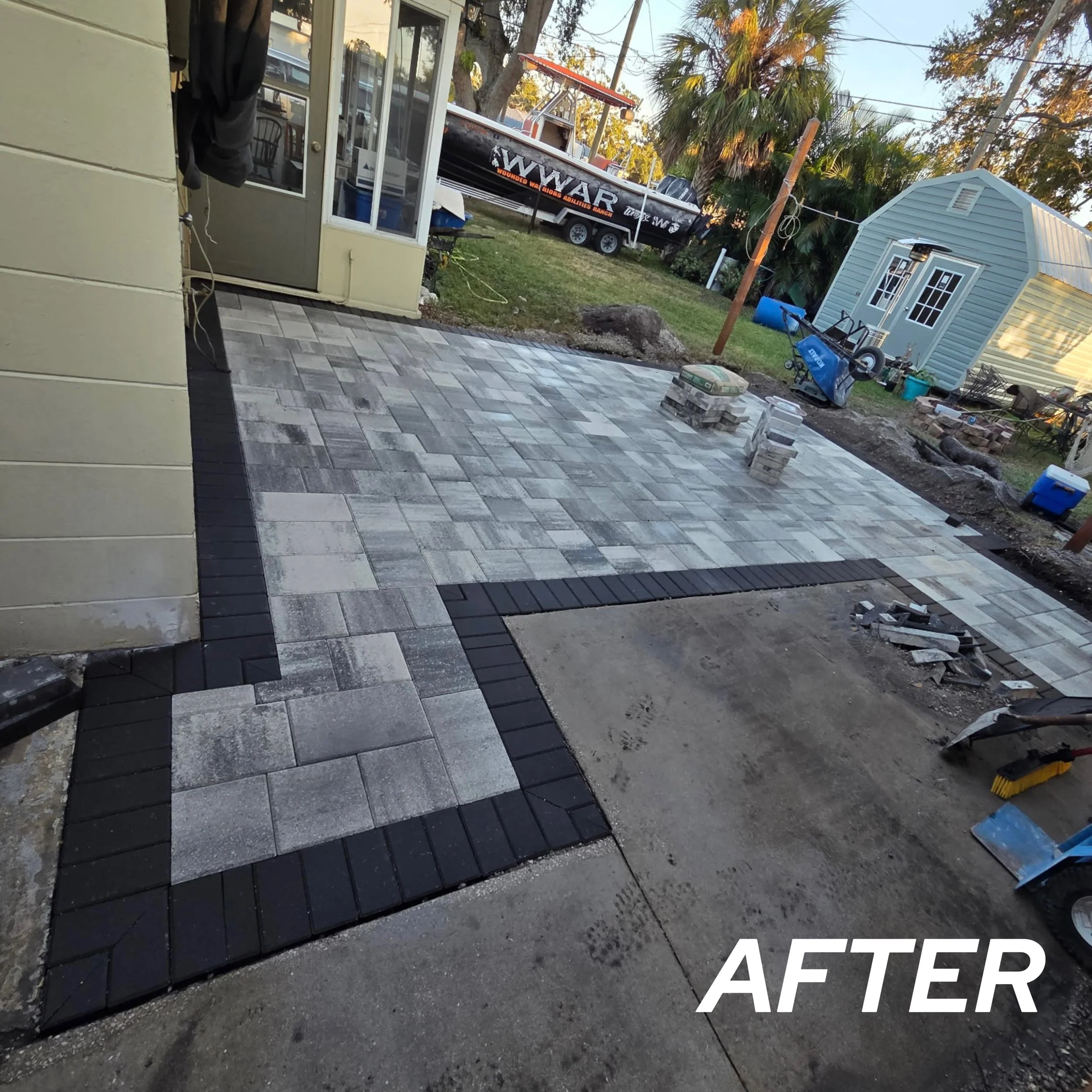 Finished outdoor patio with interlocking pavers, bordered with black bricks, and an adjacent concrete area under construction, with construction tools and materials nearby.