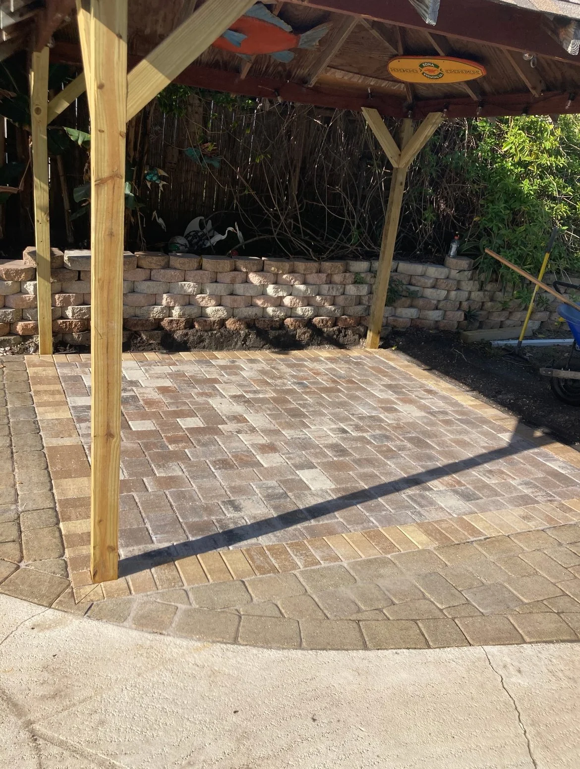 An outdoor patio with brick pavers, a wooden pergola with posts, a brick wall in the background, and gardening tools on the right side.