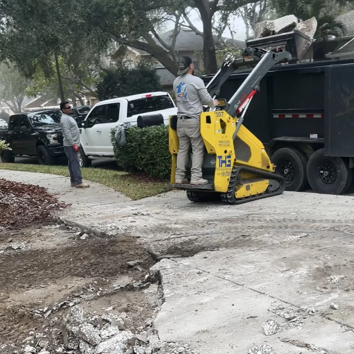 Workers repairing a damaged sidewalk next to a black trash truck using a compact track loader.