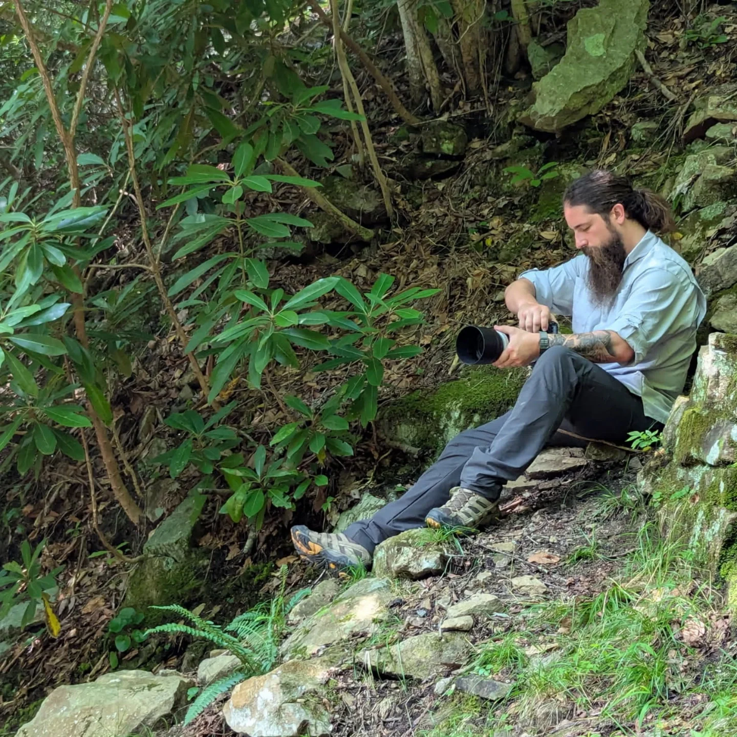 A man with a beard and long hair sitting on rocks in a forest, holding a camera lens.