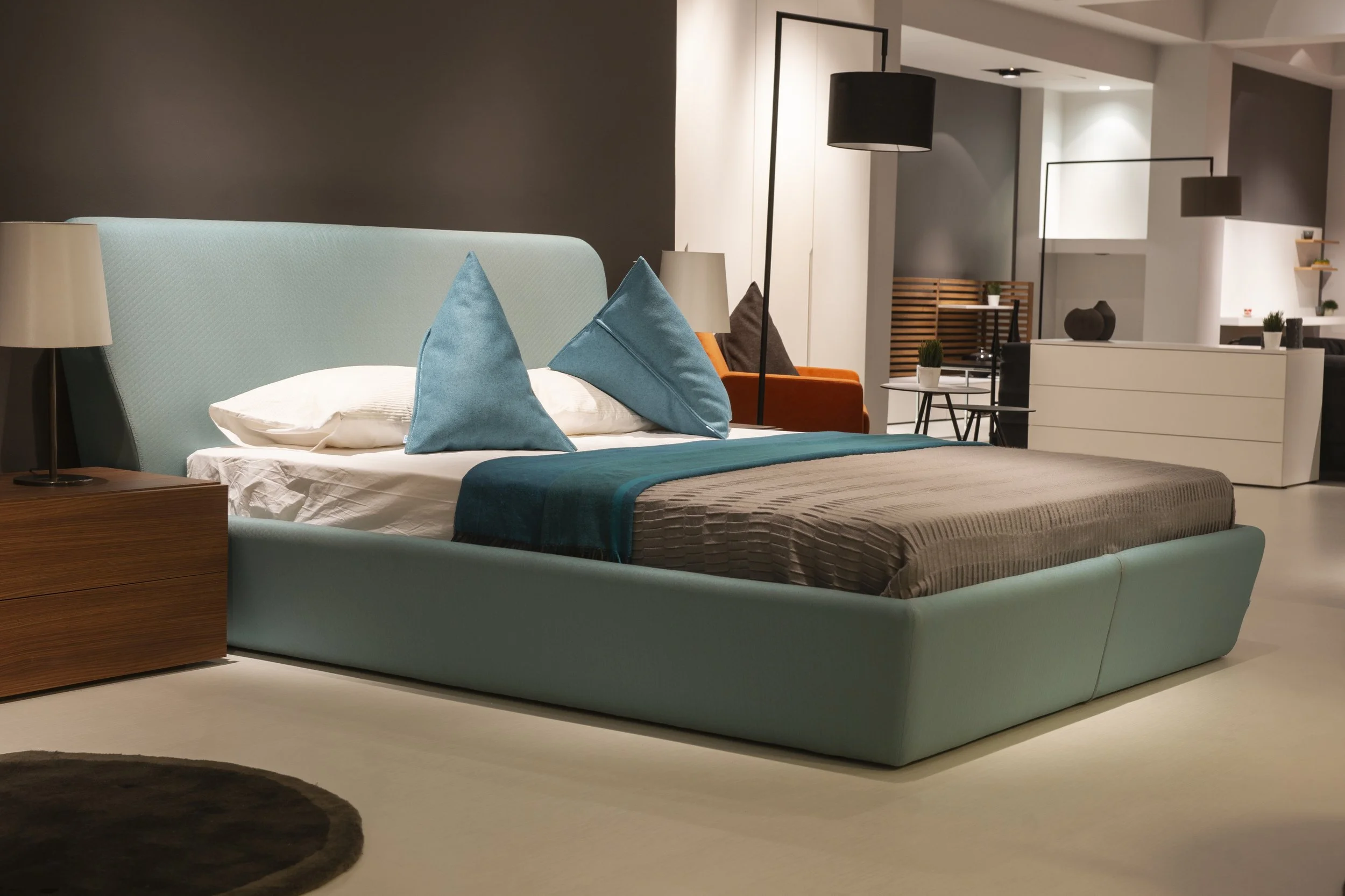 Modern bir otel odası, açık renkli yatak, mavi yastıklar ve yatak örtüsü, yan sehpa ve abajur, arka planda oturma alanı ve dekoratif objeler.
