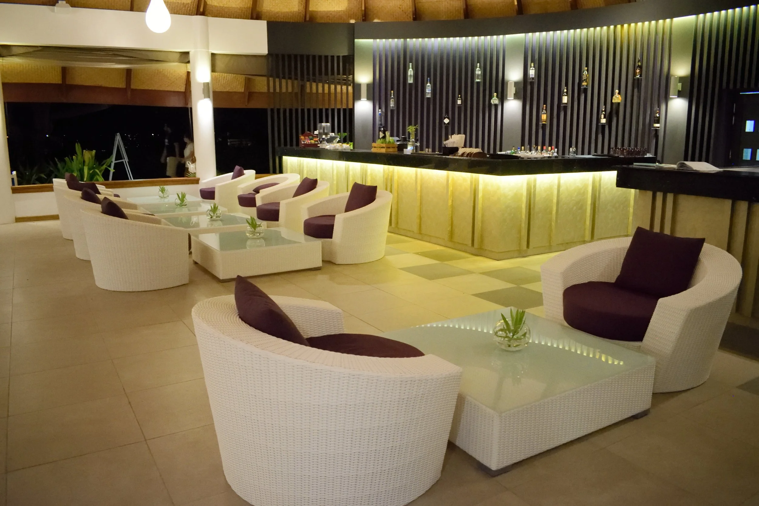 Lobi veya bar alanı, gece aydınlatmasıyla şık ve modern şekilde tasarlanmış, sandalye ve masalar, bar tezgahı ve içecekler, dekoratif ışık ve bitkilerle süslenmiş.