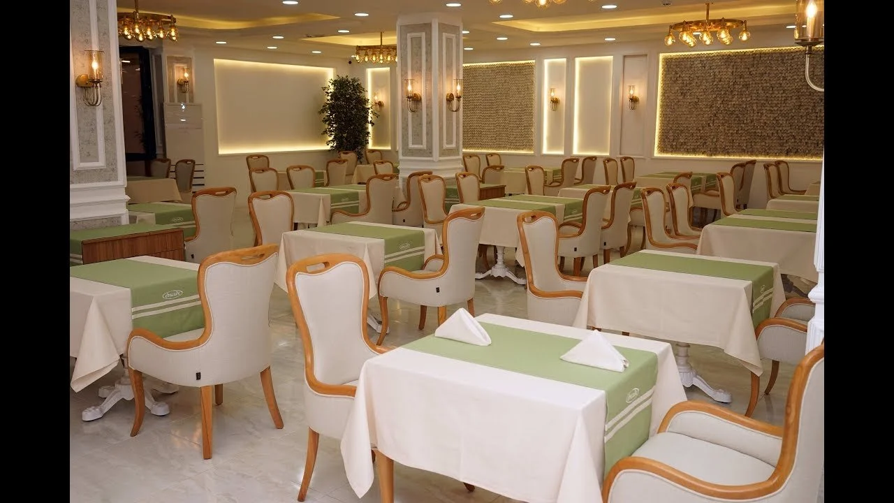 Şık ve iyi düzenlenmiş bir restoran veya yemek salonu, çok sayıda masa ve sandalye var, masa örtüleri ve peçeteler yerleştirilmiş, aydınlatma ve dekorasyon zarif. 