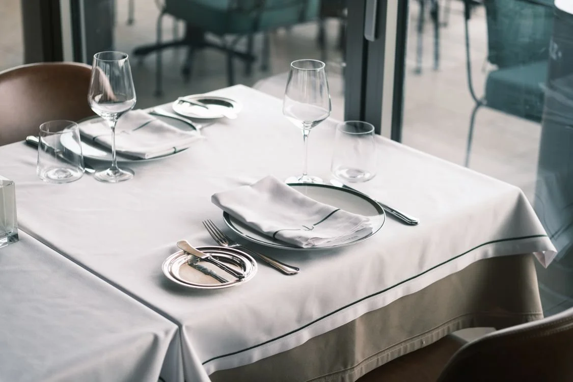 Şık bir restoran masası, üzerinde beyaz masa örtüsü, şeffaf su ve şarap bardakları, tabaklar, çatal, bıçak, kaşık ve peçete var.