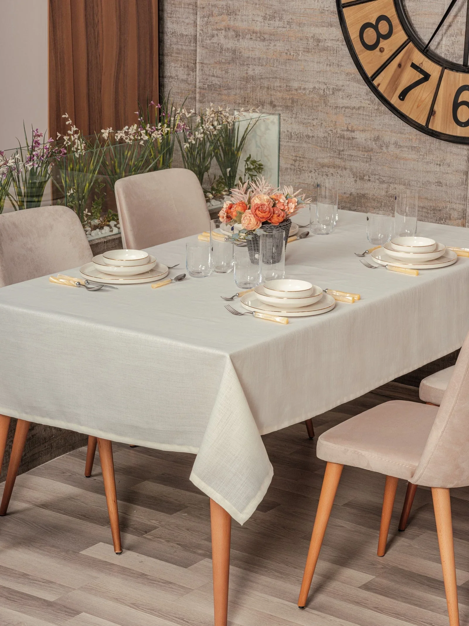 Düzenlenmiş bir yemek masası üzerinde çeşitli tabaklar, çatal, bıçak ve kaşık setleri ile birkaç boş bardak yer alıyor. Masanın ortasında narçiçeksi ve pembe tonlarında çiçeklerle süslenmiş bir buket bulunuyor. Arka planda, duvarda büyük bir saat ve birkaç bitki yer alıyor, masa ve sandalye açık bej ve ahşap tonlarında.