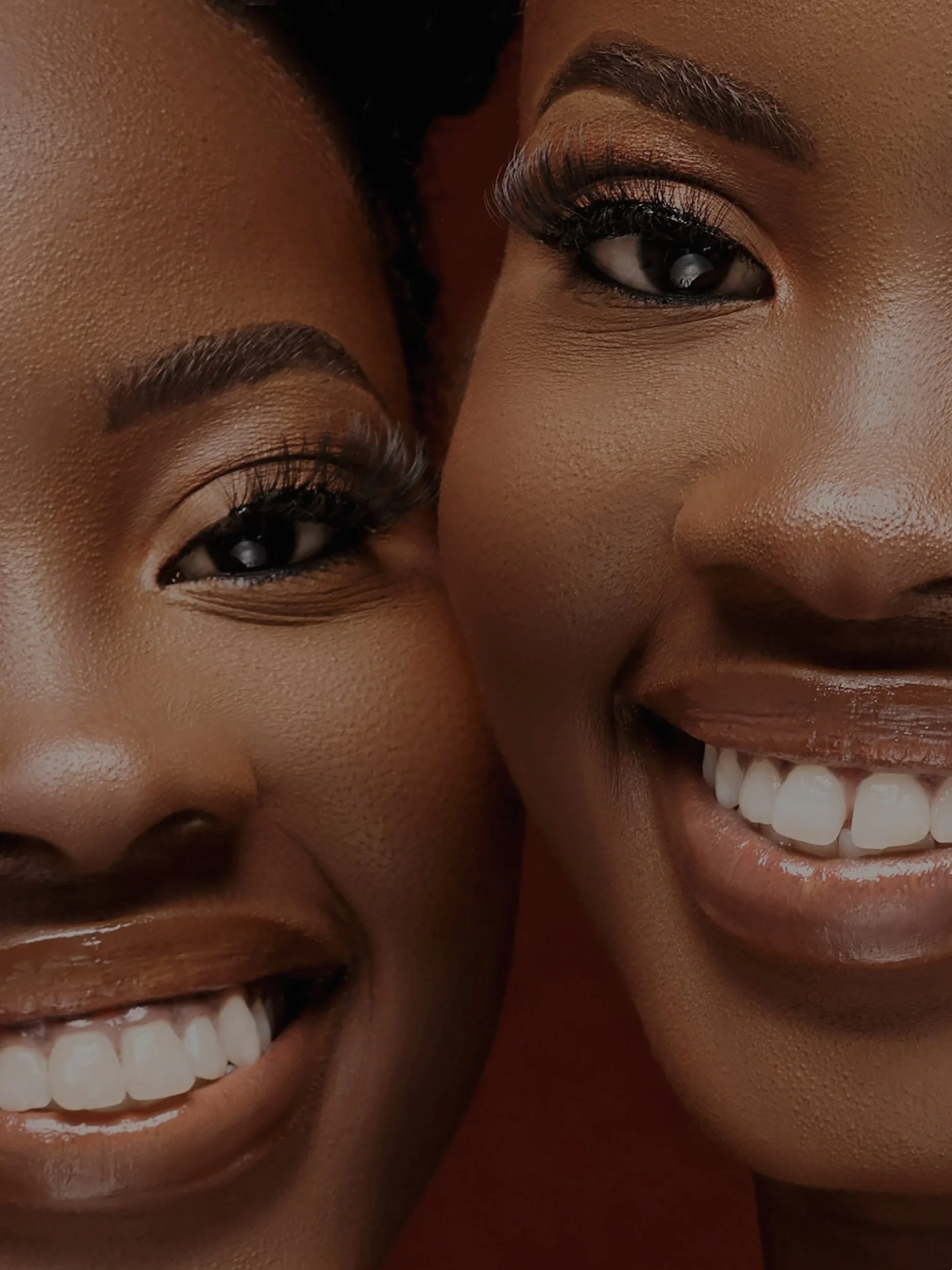 Close-up of two smiling Black women with showy teeth, makeup, and styled hair, positioned closely together.
