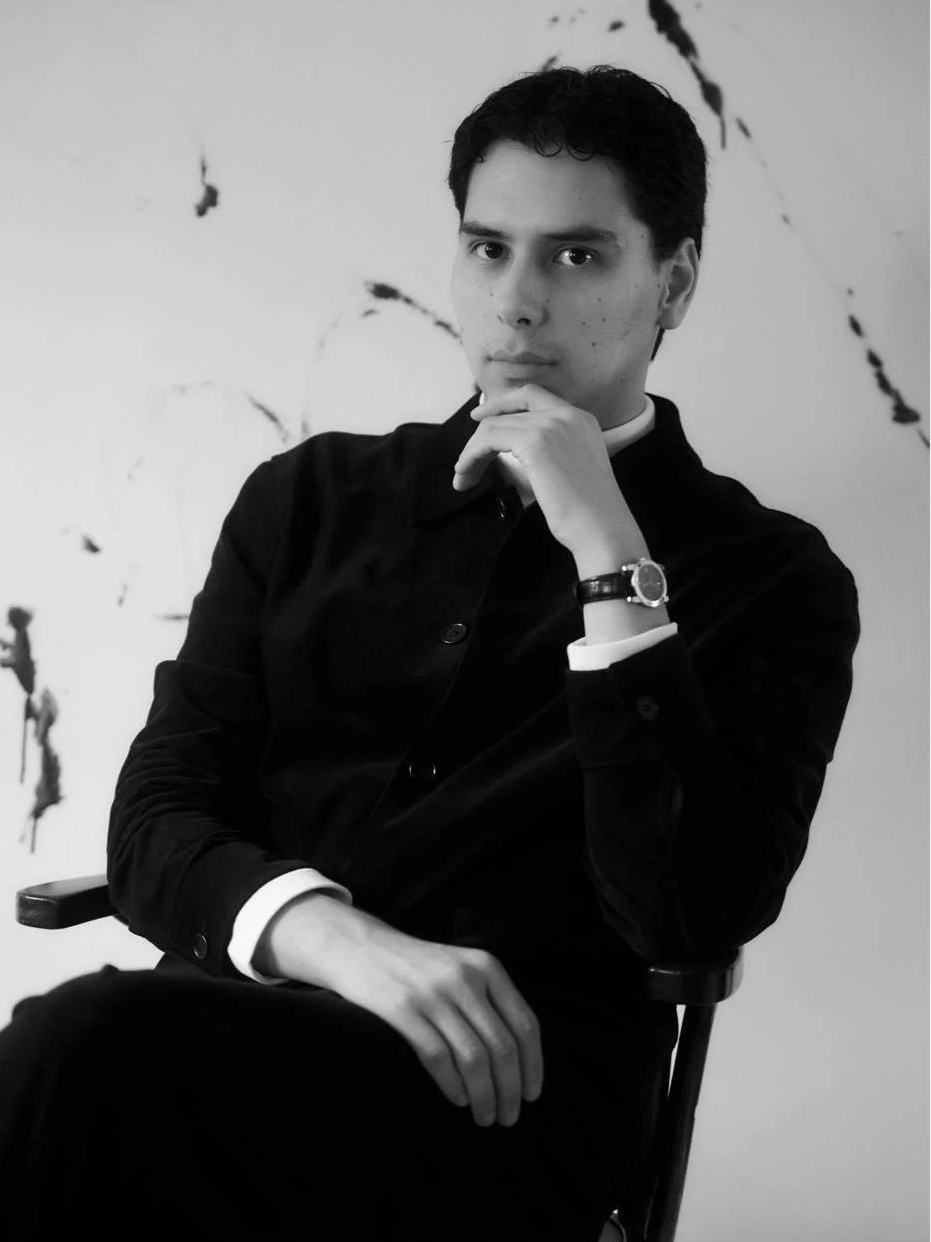 Black and white photo of a young man sitting in a chair with his chin resting on his hand, wearing a dark jacket and a wristwatch, with a serious expression, against an abstract background.