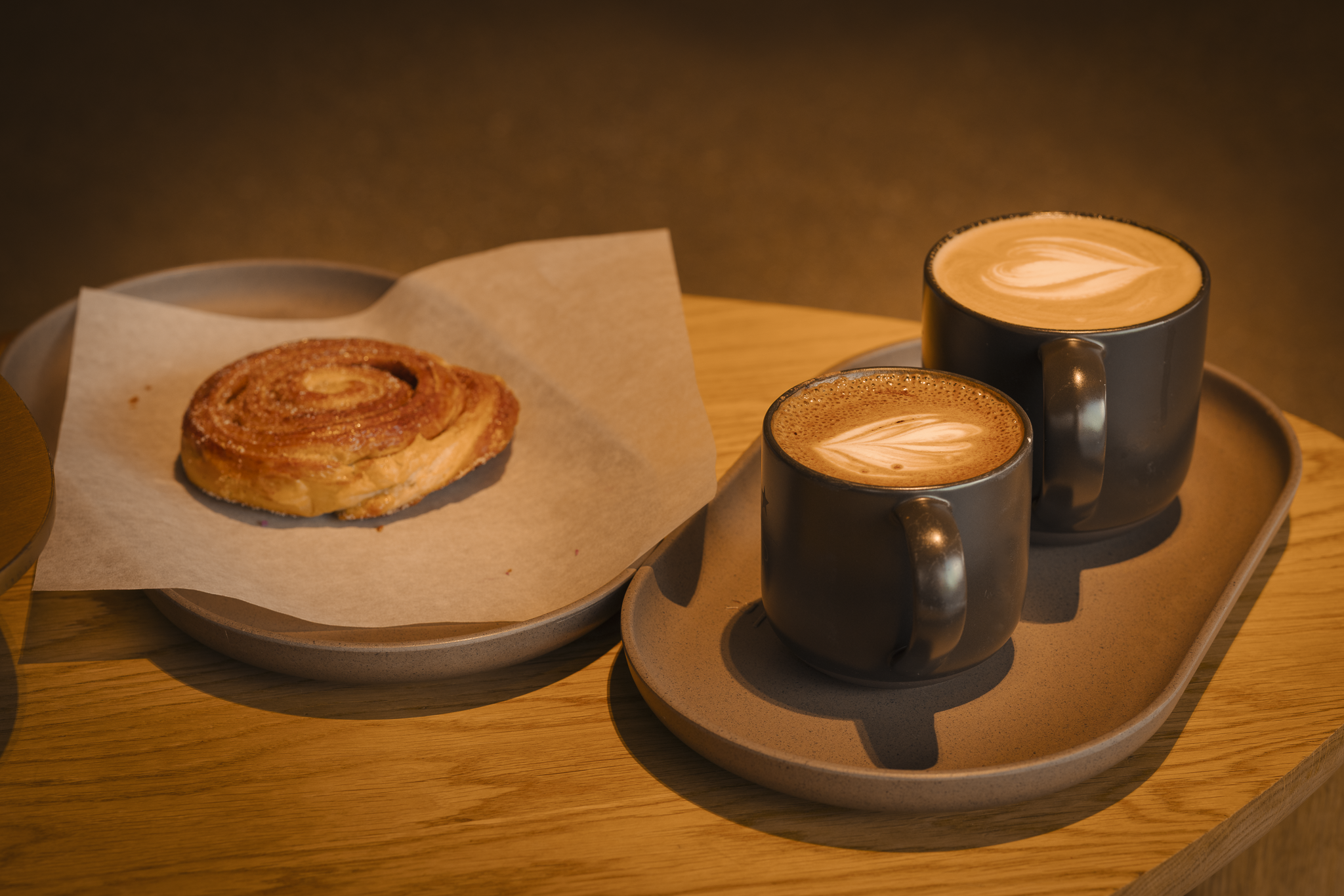 Two black coffee mugs with latte art and a cinnamon roll on a paper-lined plate on a wooden surface.