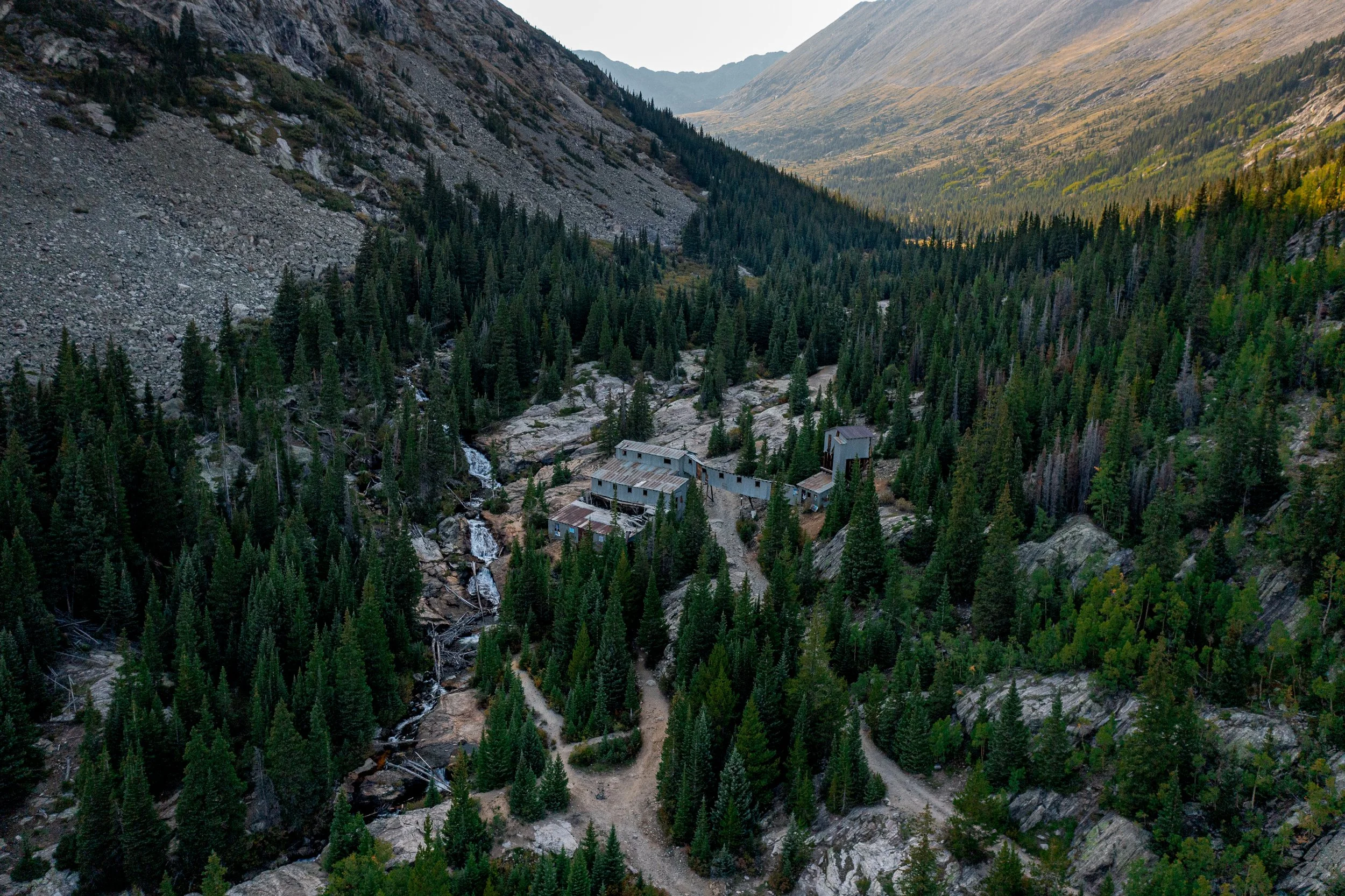 An aerial view of a mountain landscape with dense evergreen trees and a few old wooden structures near a small creek.