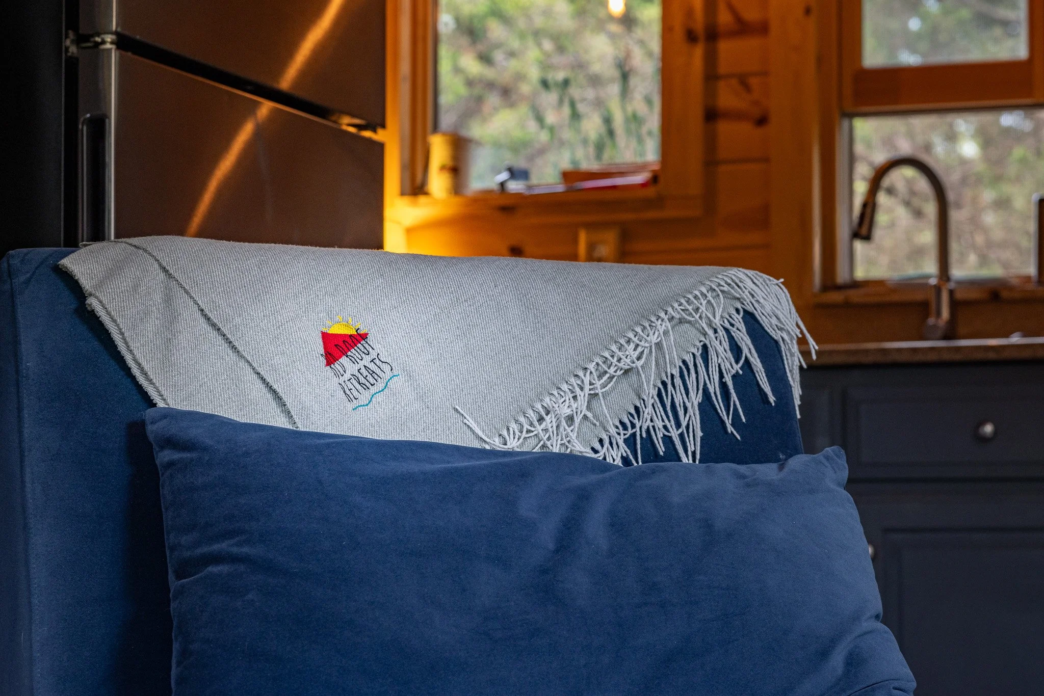 A cozy interior scene featuring a blue couch with a beige throw blanket embroidered with a sunset and the words 'RETREAT' draped over the back. In the background, there are wooden walls, a window showing trees outside, and a sink faucet.