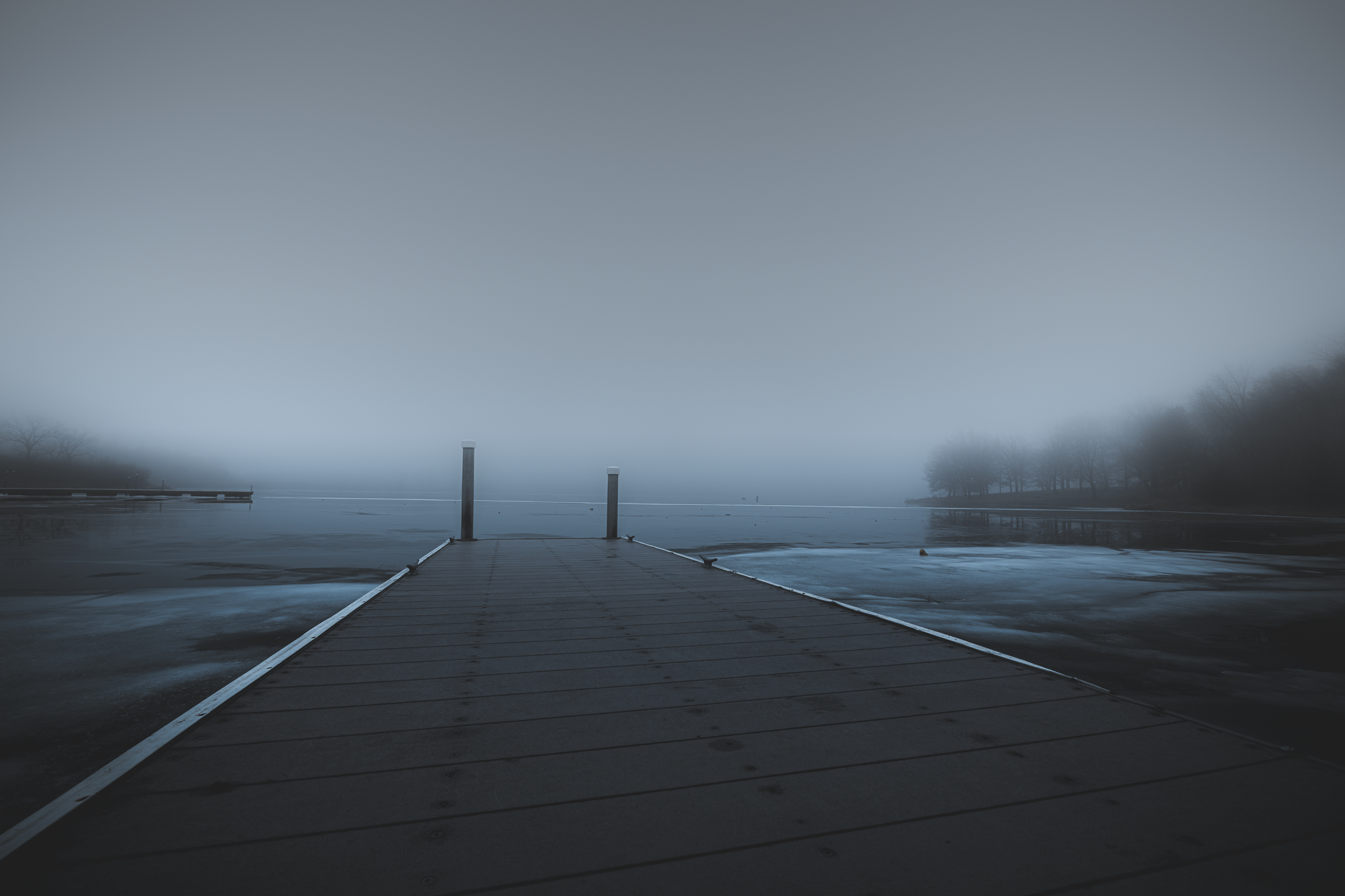 A foggy lakeside scene with a wooden dock extending into the water, surrounded by dense fog and silhouettes of trees in the distance.