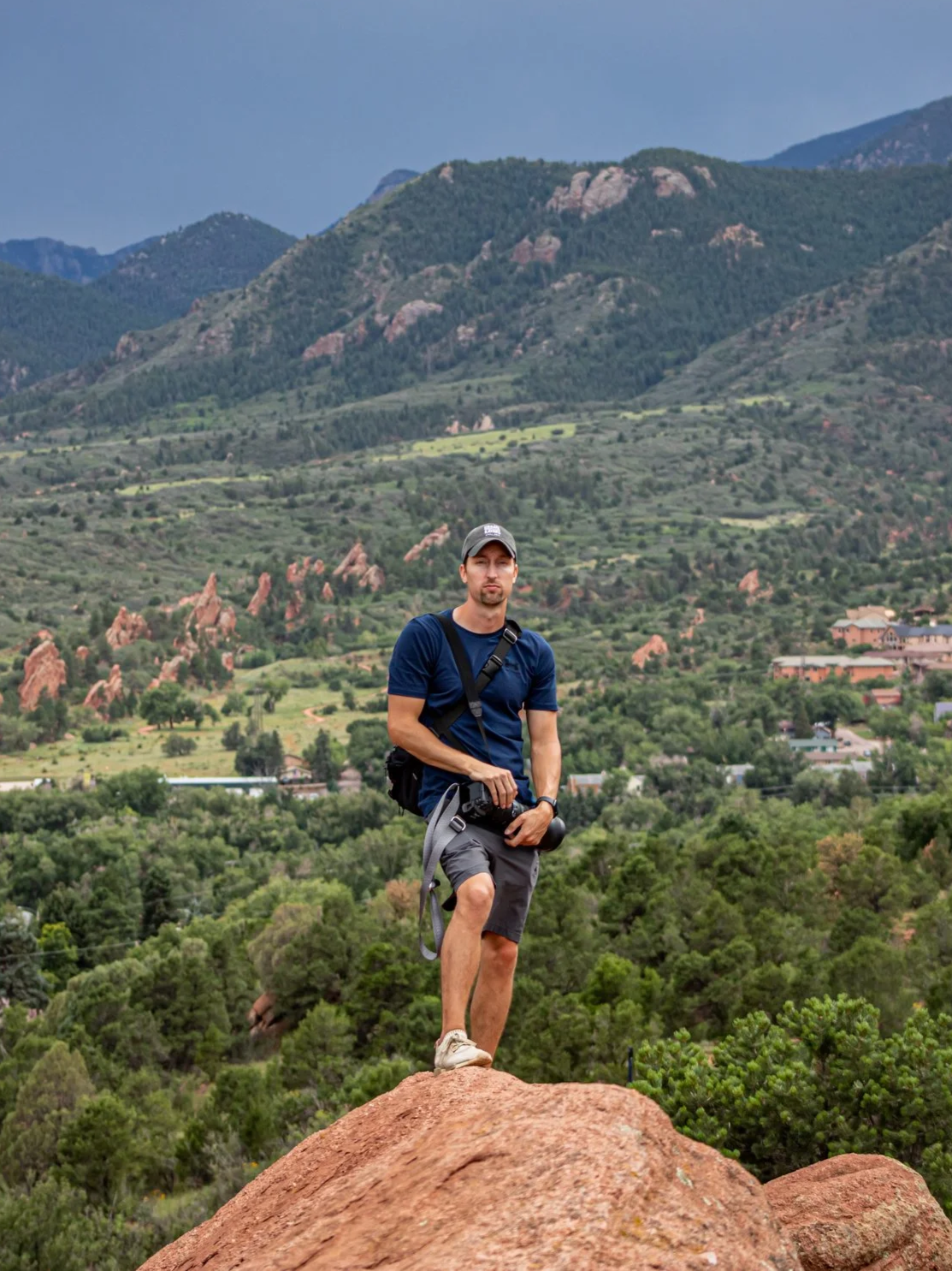 Man standing on a large rock with mountains and greenery in the background.