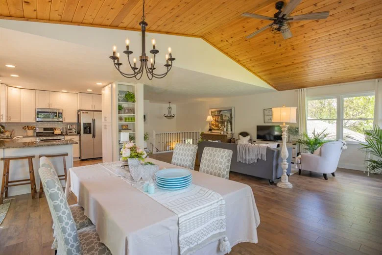Open-concept living and dining area with a wooden ceiling, chandelier, dining table with chairs, sofa, armchair, lamp, TV, and large windows, sunlight streaming in.