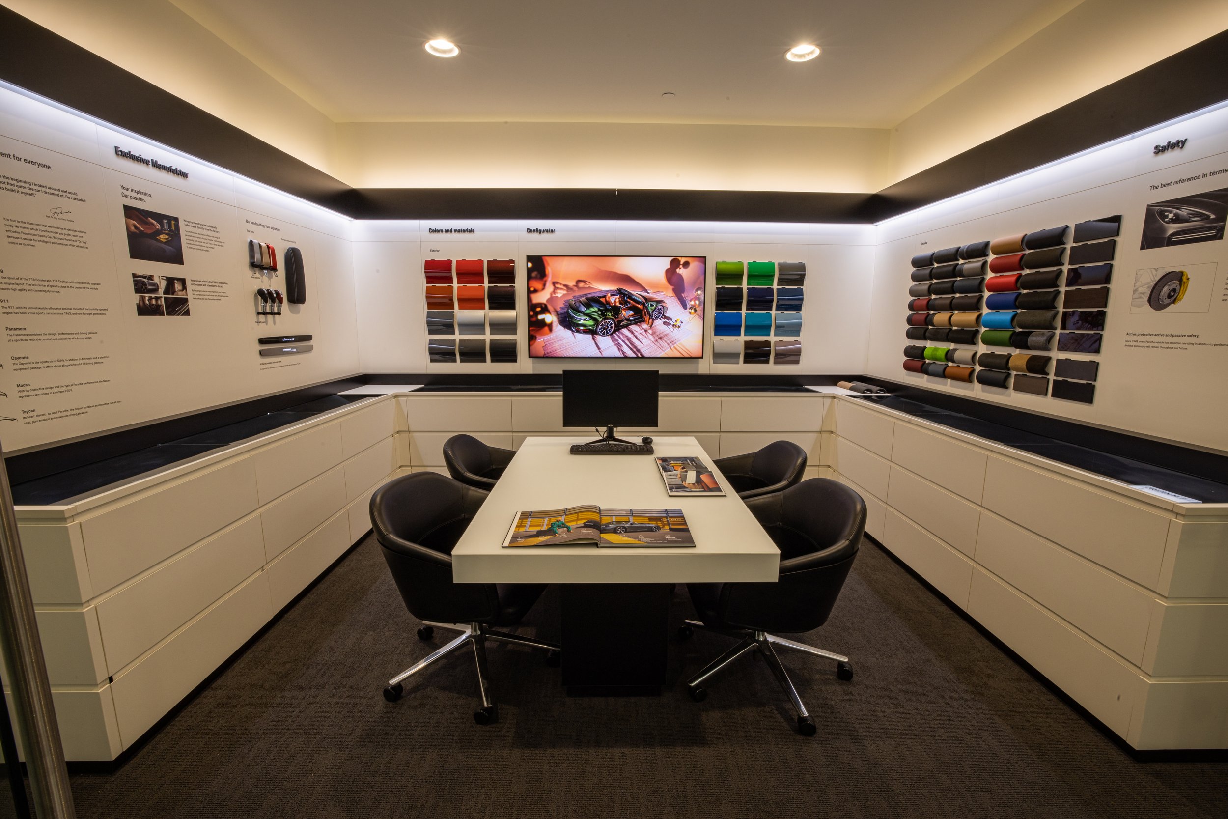 A car customization room featuring paint color samples arranged on the walls, a central desk with a monitor and brochures, and chairs around the desk.