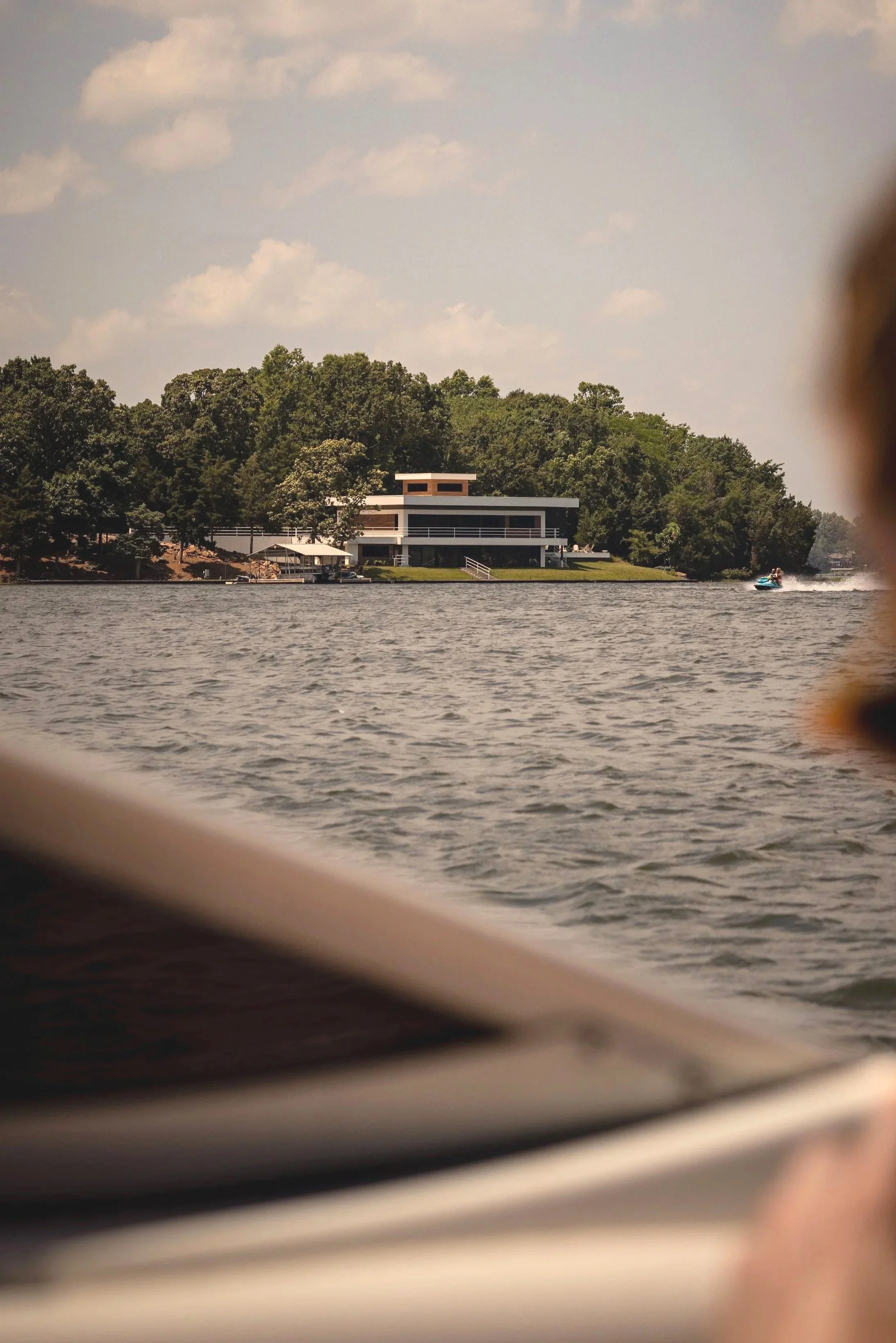 View of a modern house on a wooded shoreline across a lake, partially obscured by boat parts in the foreground, with a boat speeding past on the right.