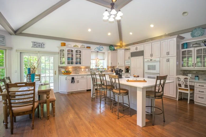 Kitchen with white cabinets, a central island with seating, hardwood floors, and a dining table with chairs. Decor includes blue and green accents and the word 'Eat' on the wall.