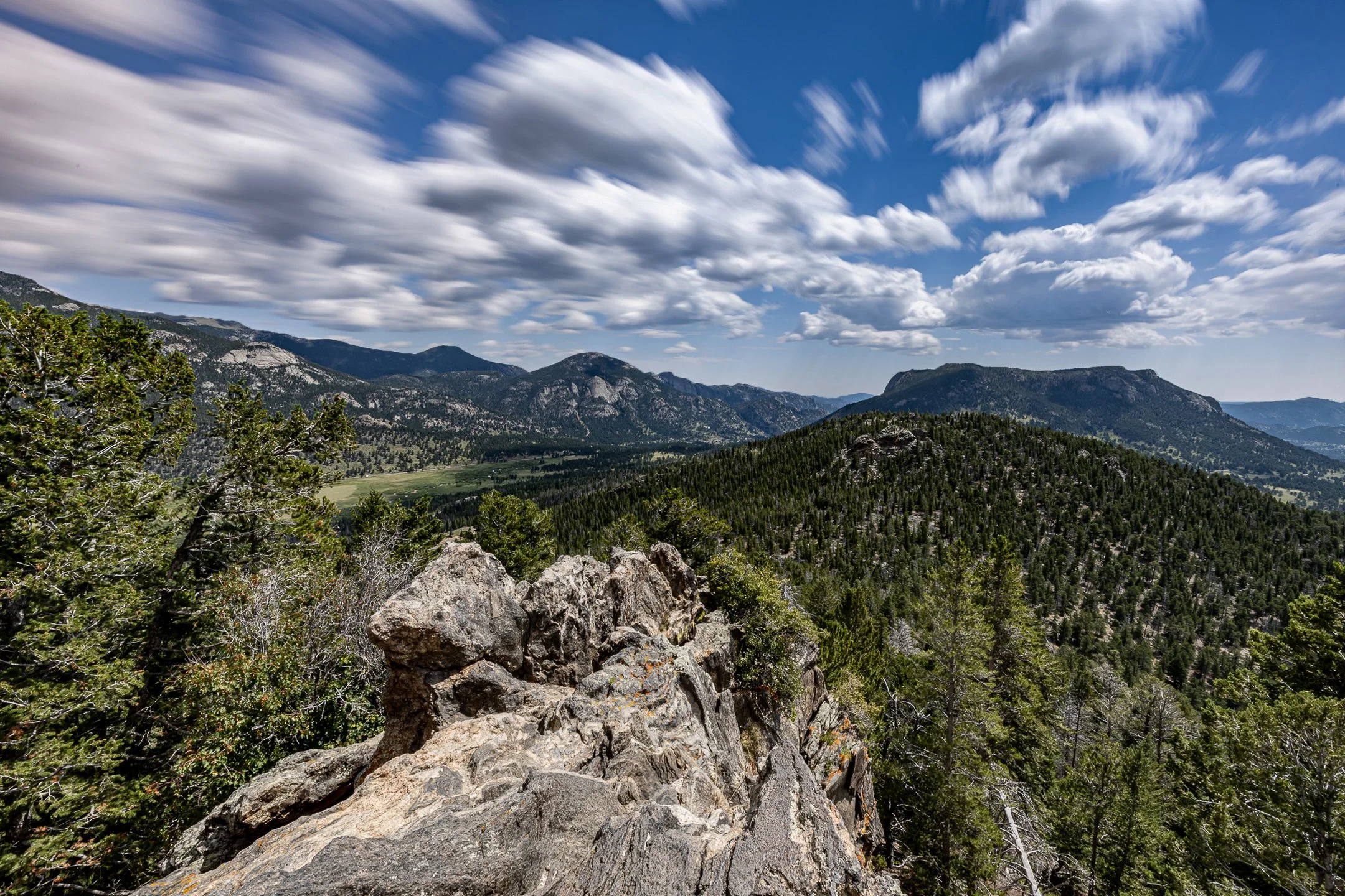 A mountainous landscape with a partly cloudy sky, dense evergreen forests covering the hills, and rocky outcroppings in the foreground.