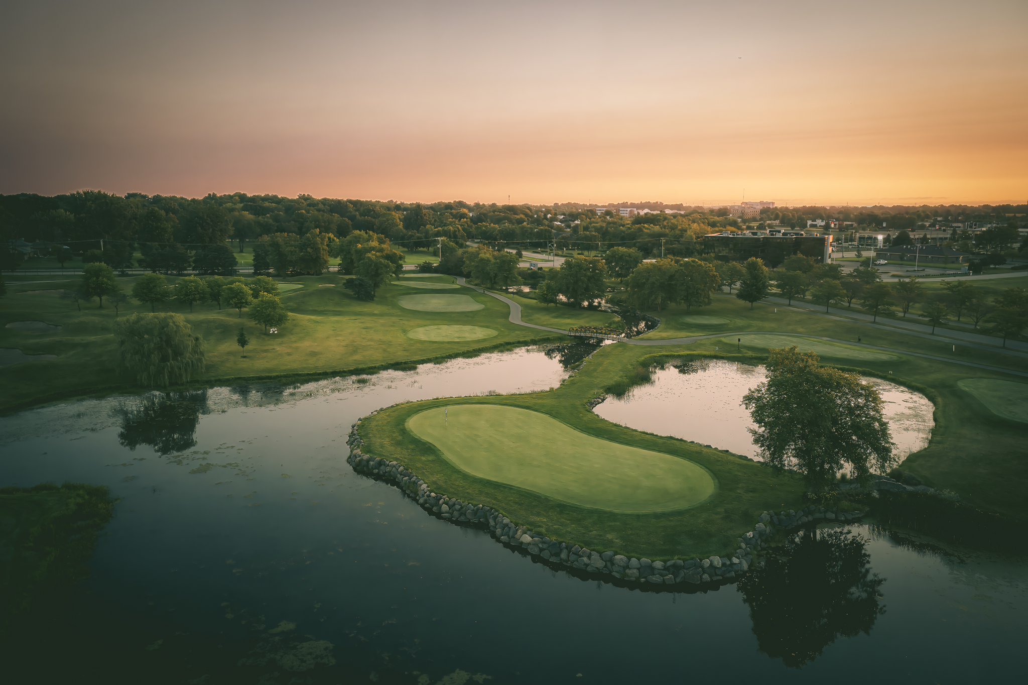 Aerial view of a golf course at sunset, featuring a green surrounded by water with trees and pathways.