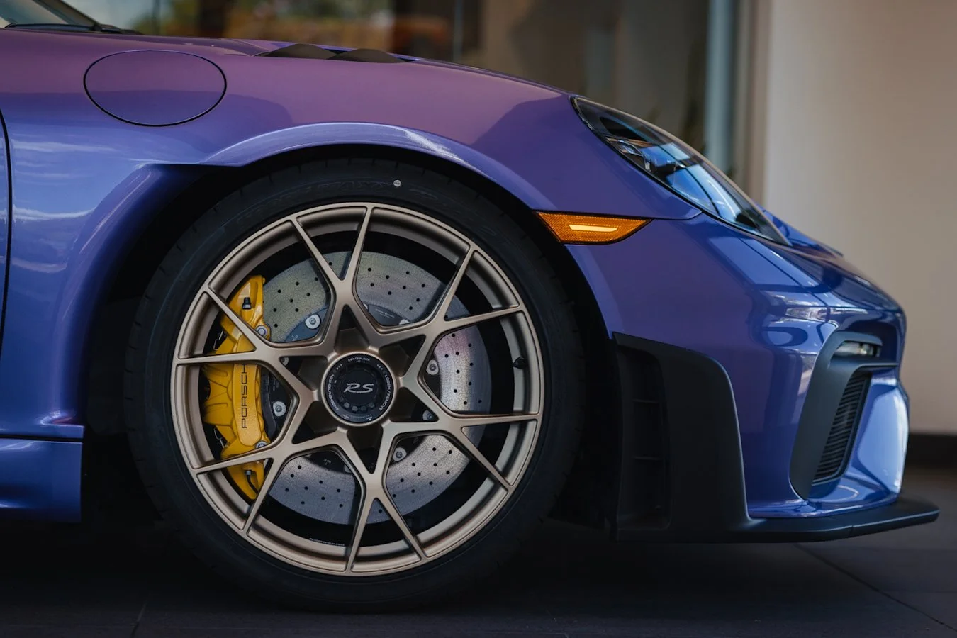 Close-up of a blue Porsche sports car's front left side, showing a large gold multi-spoke wheel with a yellow brake caliper and drilled brake disc.
