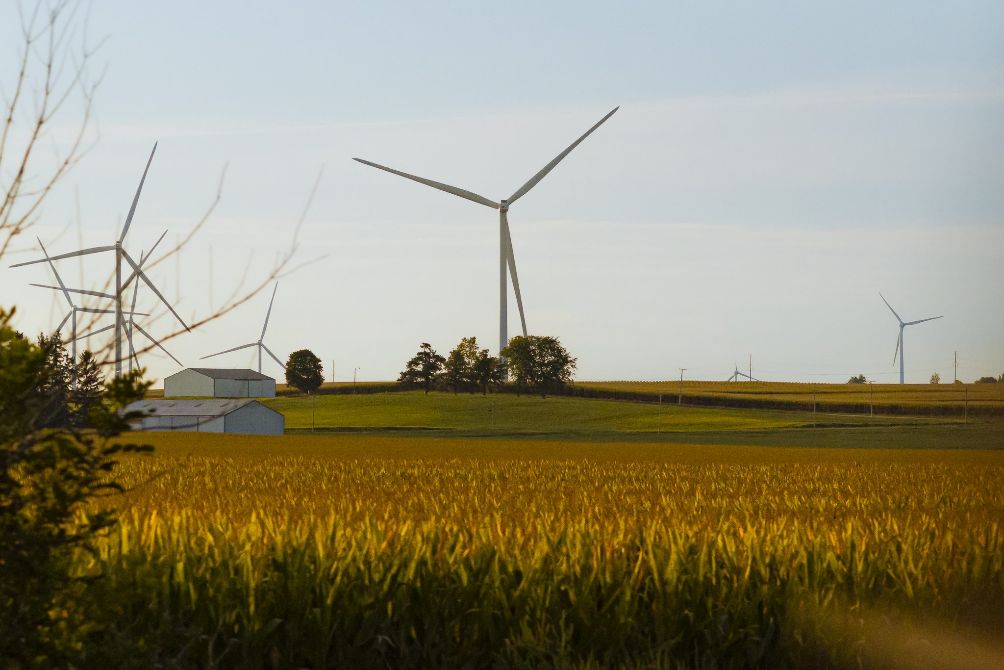 A rural landscape with wind turbines in a field, farm buildings, trees, and a sky with scattered clouds.