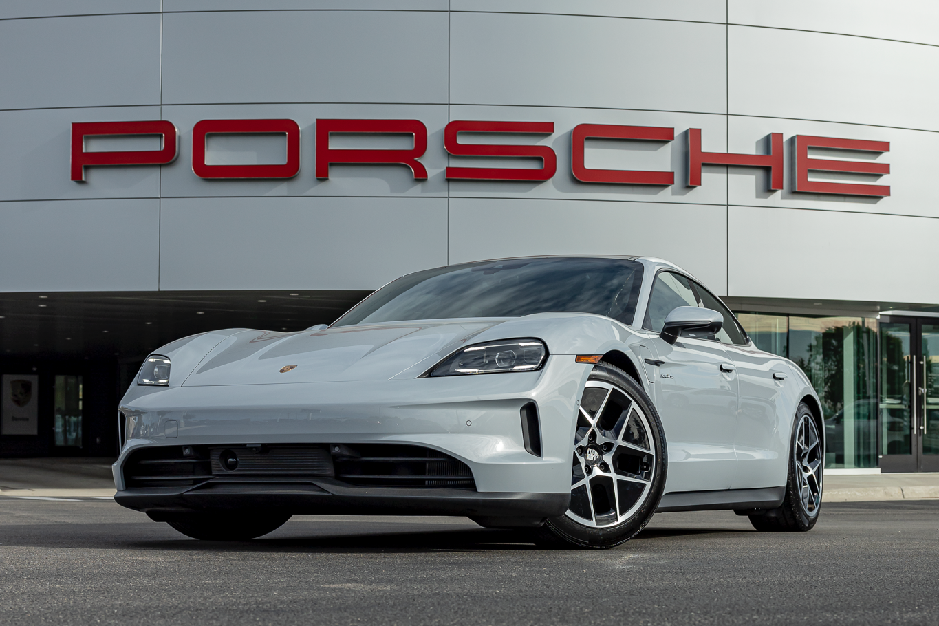 A white Porsche sports car parked in front of a Porsche dealership with a large red Porsche sign on the building.