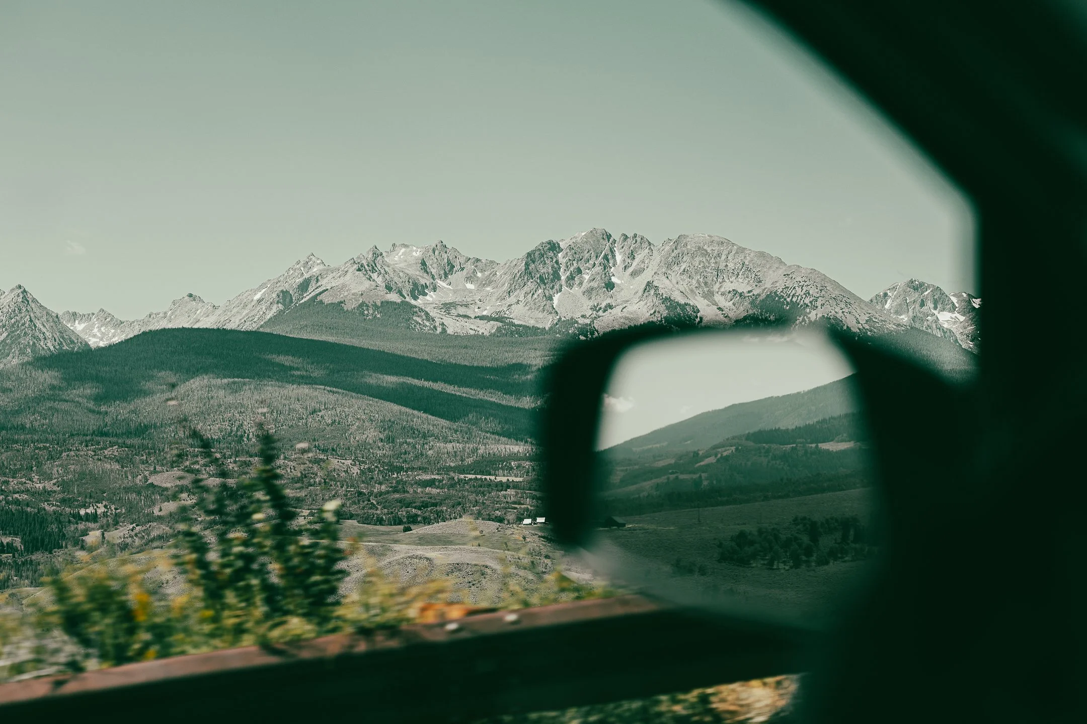 Mountain landscape with snowy peaks viewed through a car's side mirror and window.