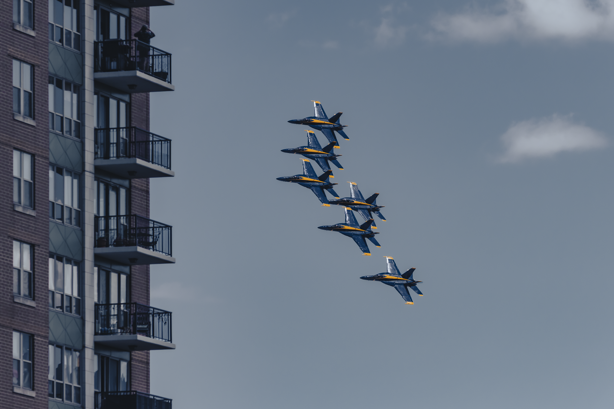 Six fighter jets flying in a tight diamond formation near a residential building with balconies, against a blue sky with some clouds.