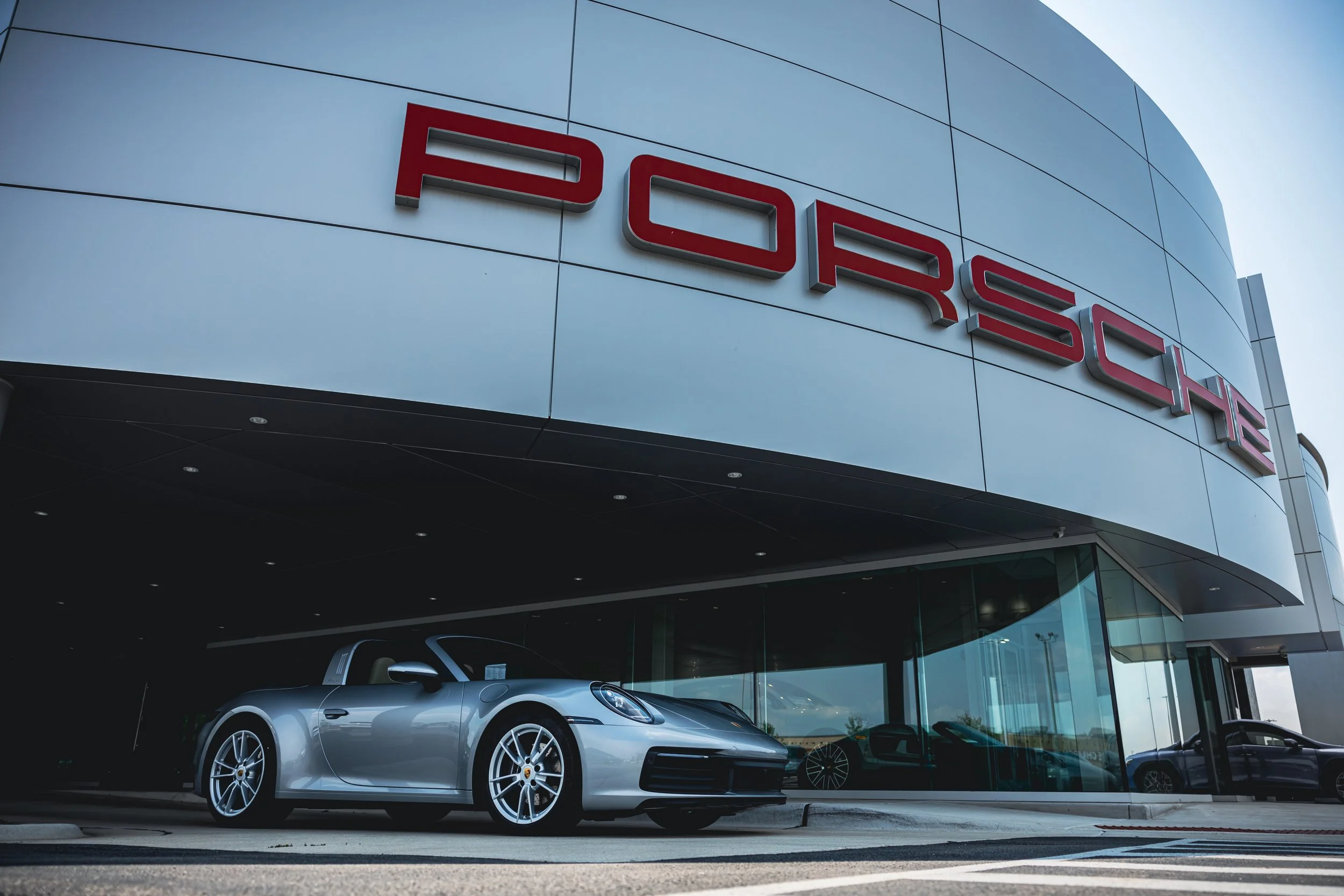 A silver Porsche sports car parked outside a Porsche dealership building with the red Porsche logo.