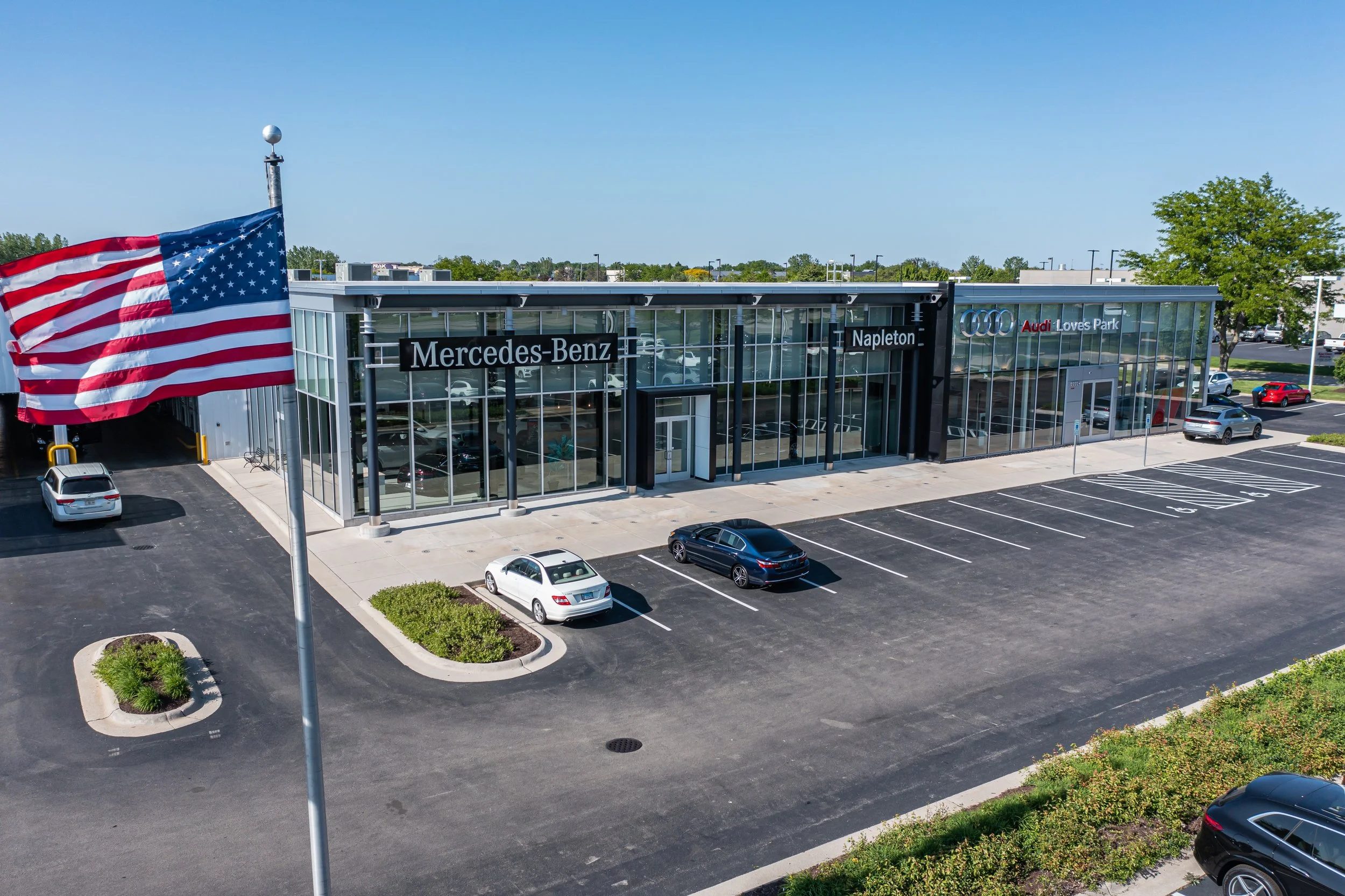 An aerial view of a modern car dealership with glass storefronts labeled Mercedes-Benz, Napleton, and Audi Loves Park, and a large American flag. Parking lot in front with a few cars and landscaped islands.