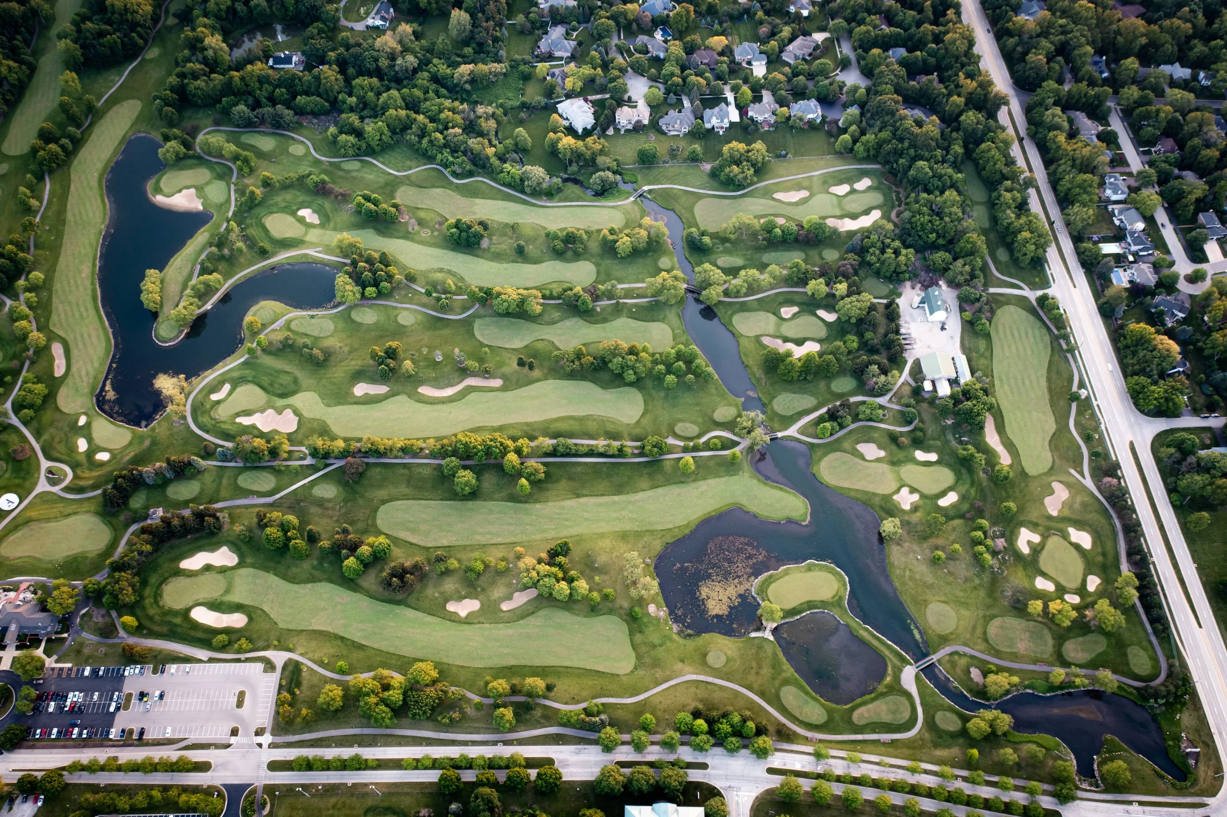 Aerial view of a golf course with several holes, water hazards, sand traps, and surrounding trees, bordered by residential neighborhoods and a main road.