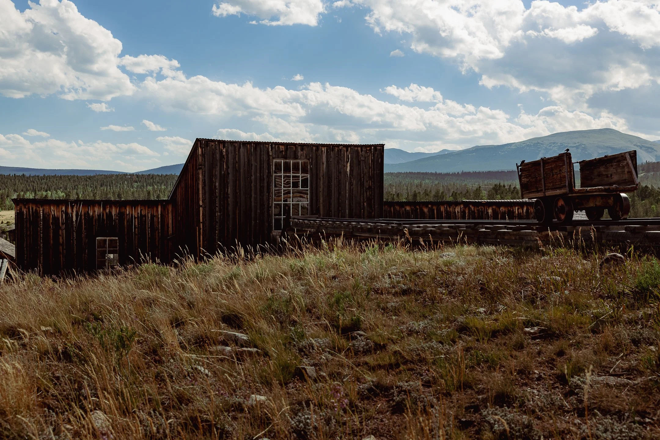 An old, weathered wooden building with metal and glass windows situated on grassy terrain, with a railway track and cart nearby, against a backdrop of mountains and a partly cloudy sky.
