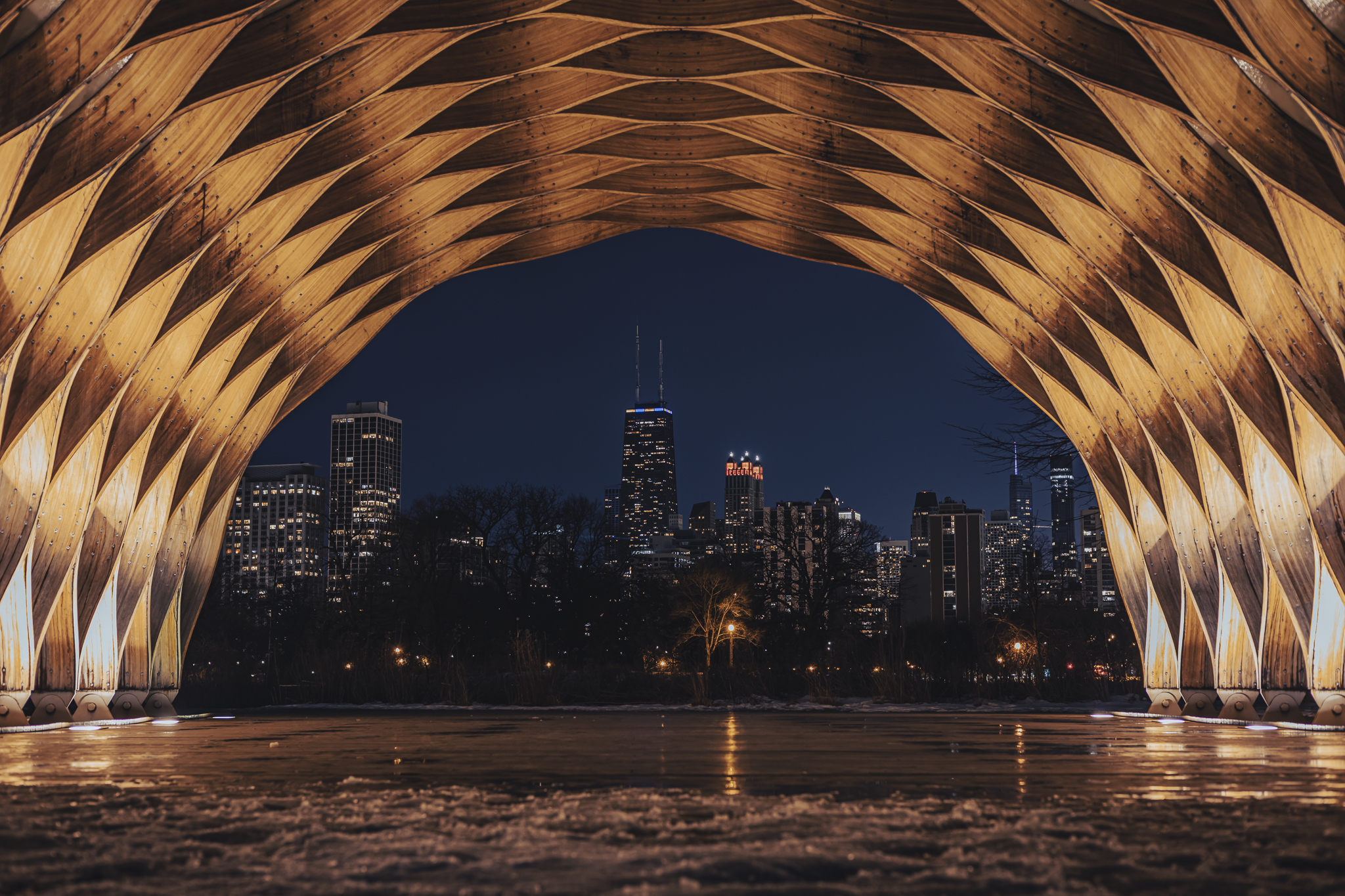 Night view of Chicago skyline seen through the wooden arches of a bridge or pavilion with the John Hancock Center prominent in the background.