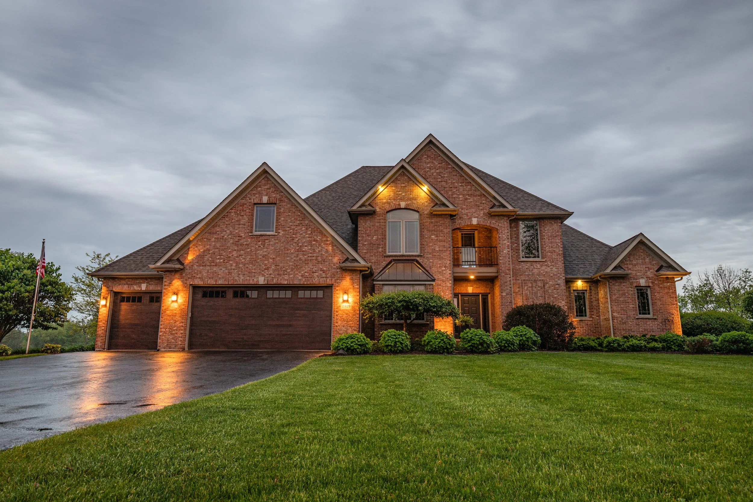 A large brick house with a three-car garage, lit up exterior lights, a well-manicured lawn, and a cloudy sky in the background.