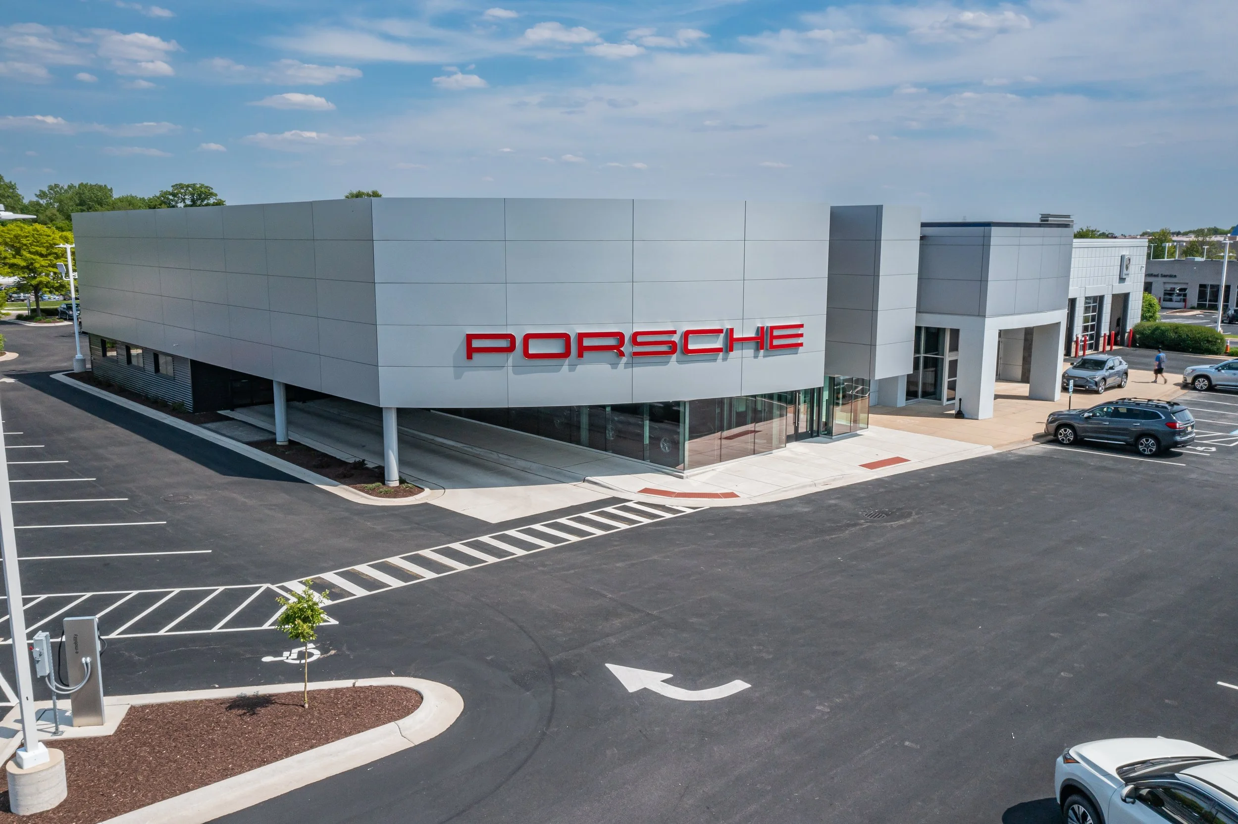 An aerial view of a modern Porsche car dealership building with a large red Porsche logo on the front, surrounded by a parking lot with a few cars parked and a clear, partly cloudy sky overhead.