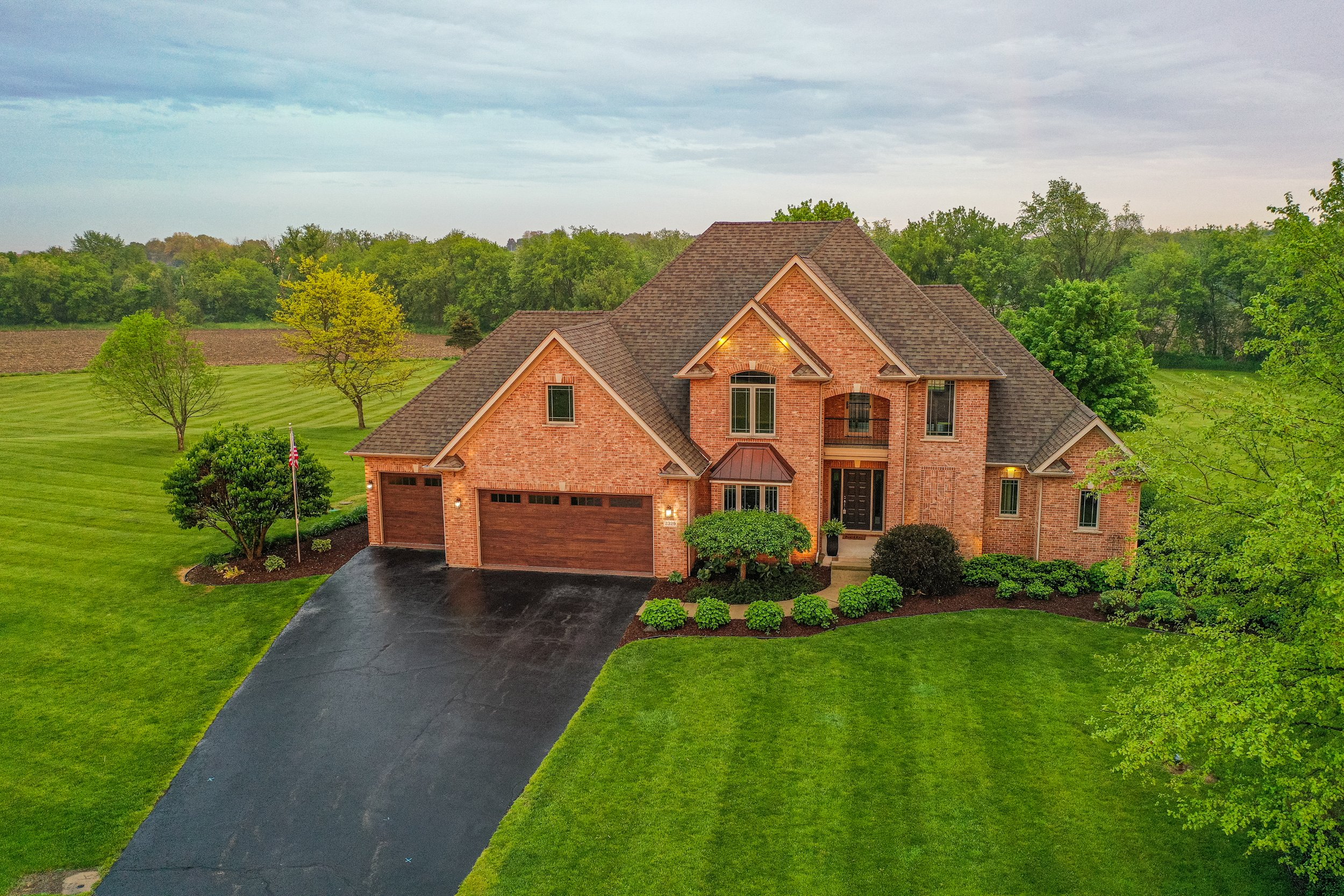 An aerial view of a large brick house with a dark shingled roof, a three-car garage, surrounded by green lawns and trees, during dusk with outdoor lighting turned on.