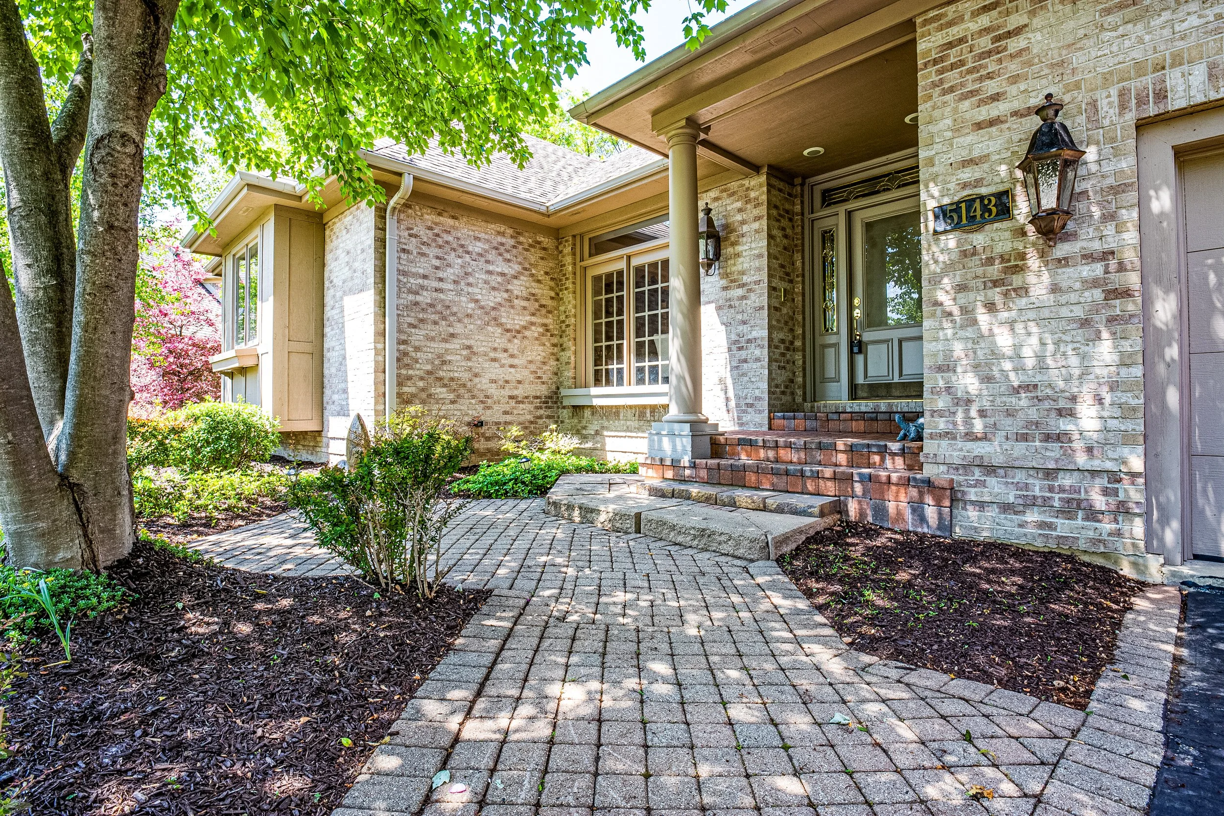 Front yard of a house with a brick walkway, stairs, and a porch, surrounded by trees and bushes, with house number 5143 on the wall.