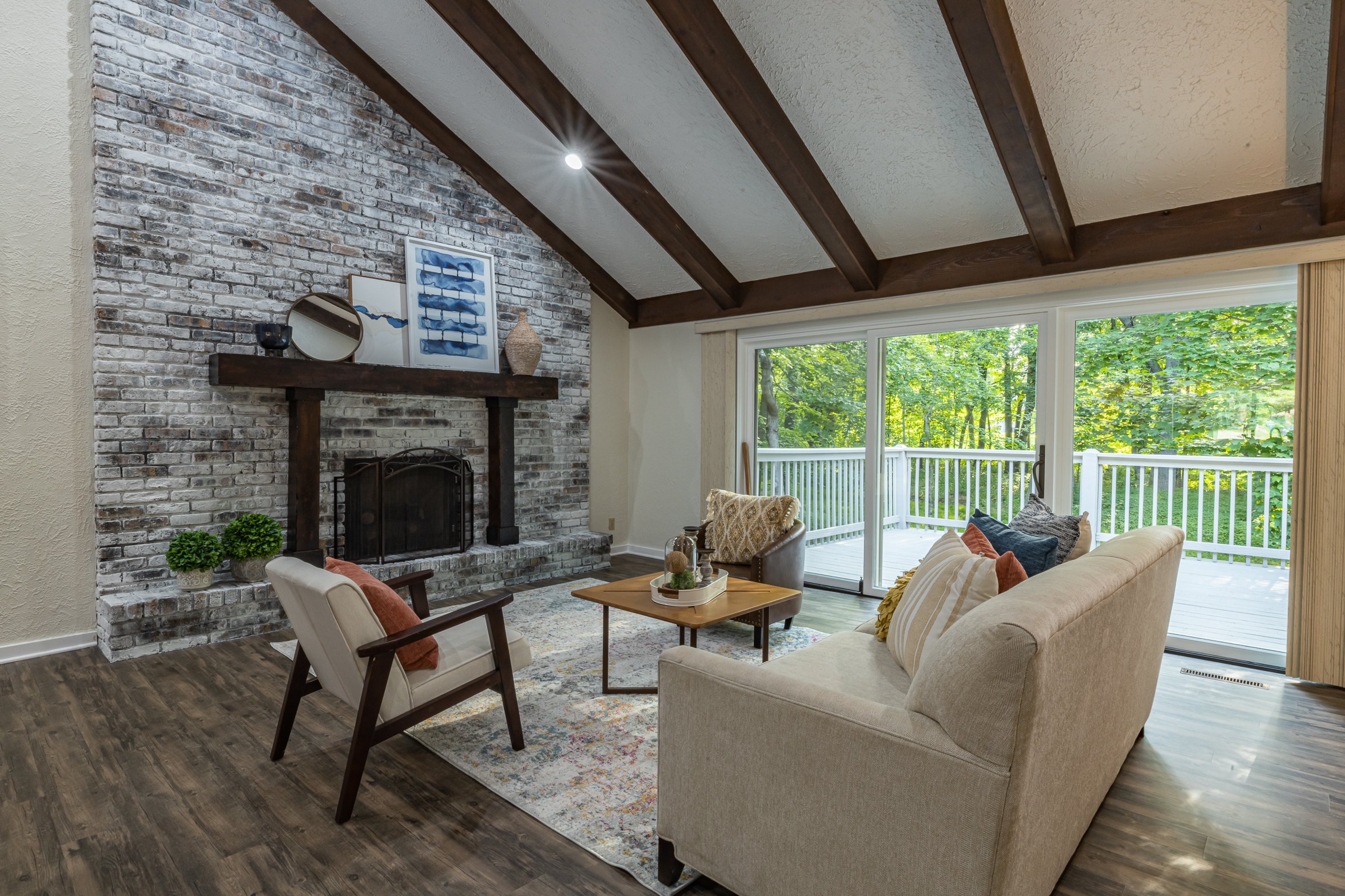 Living room with large sliding glass doors overlooking a wooded backyard, a beige sofa, an armchair, a wooden coffee table, a decorative rug, a brick fireplace with a dark wooden mantel, and wall art with blue accents.