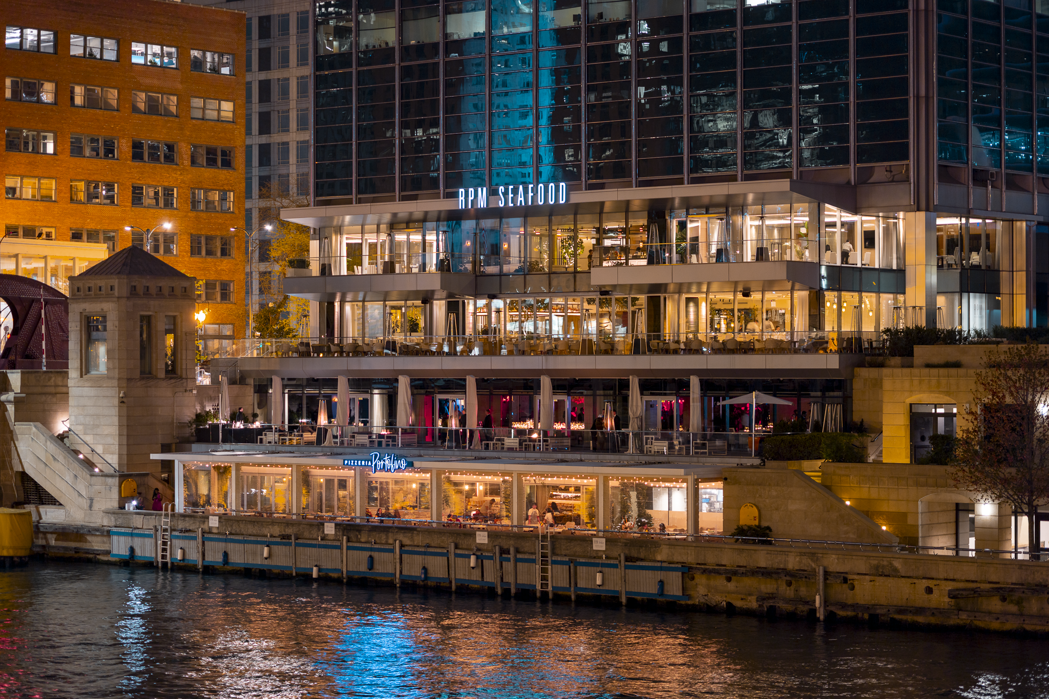 Night view of a modern multi-story waterfront restaurant building with illuminated interior and exterior lights, the sign reads 'RPM SEAFOOD' on the top level, and 'Pizzare Portofino' on the lower level, with water in the foreground reflecting the li