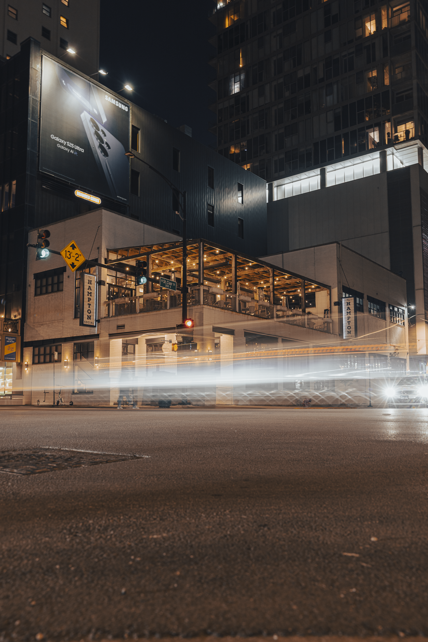 Nighttime city street scene featuring a building with illuminated restaurant outdoor seating, streetlights, blurred vehicle lights, traffic signals, and billboards.