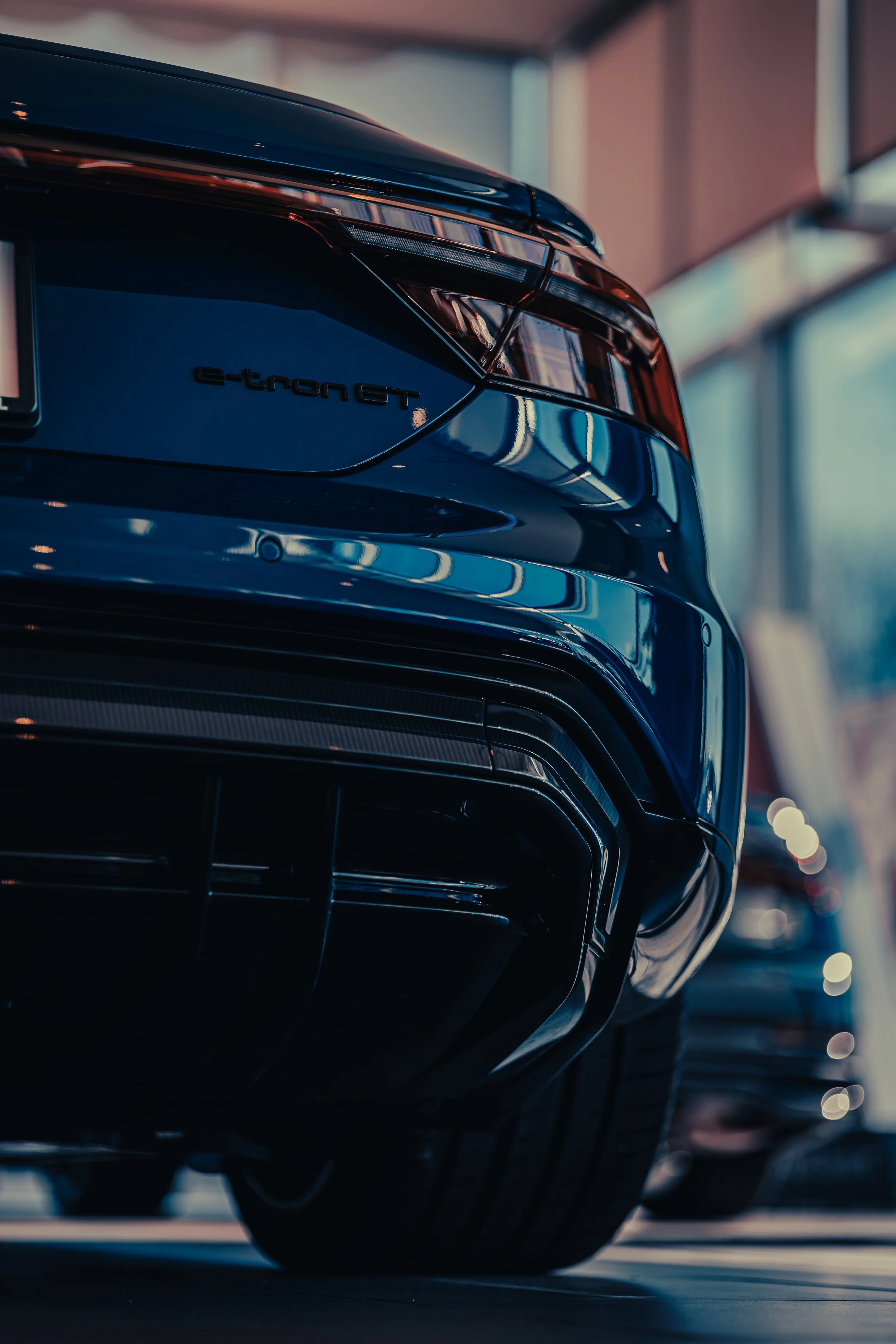 Close-up of the rear end of a dark blue Audi e-tron GT electric vehicle, showing tail light, rear bumper, and tire in a showroom.