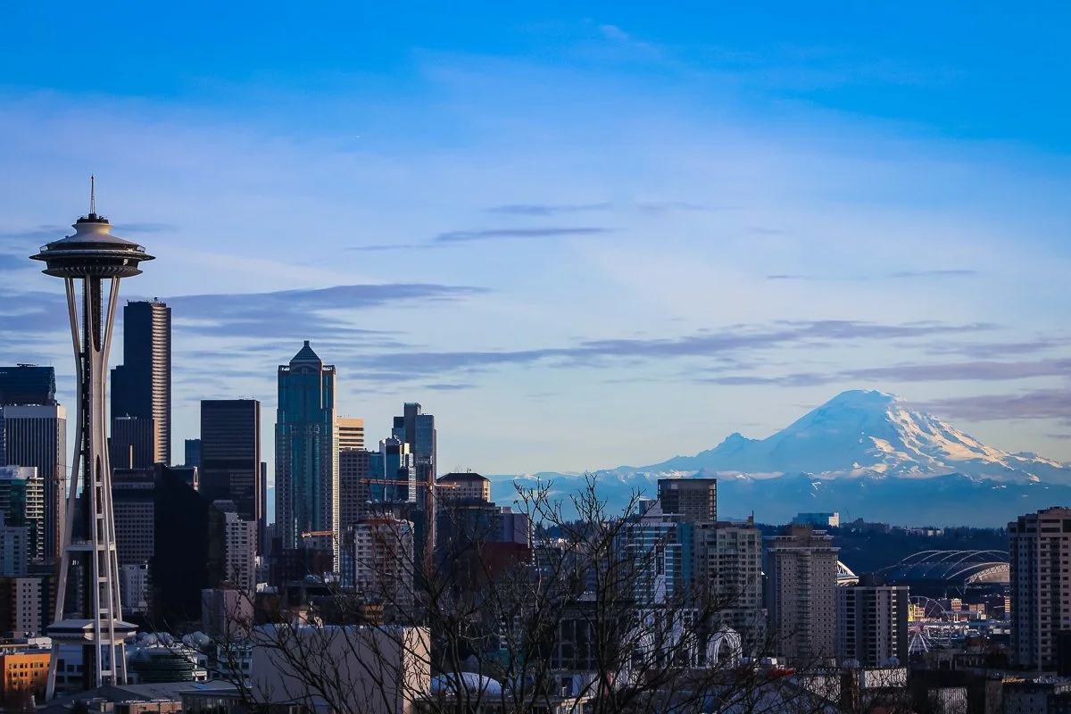 Skyline of Seattle with the Space Needle and downtown buildings, and Mount Rainier in the distance under a partly cloudy blue sky.