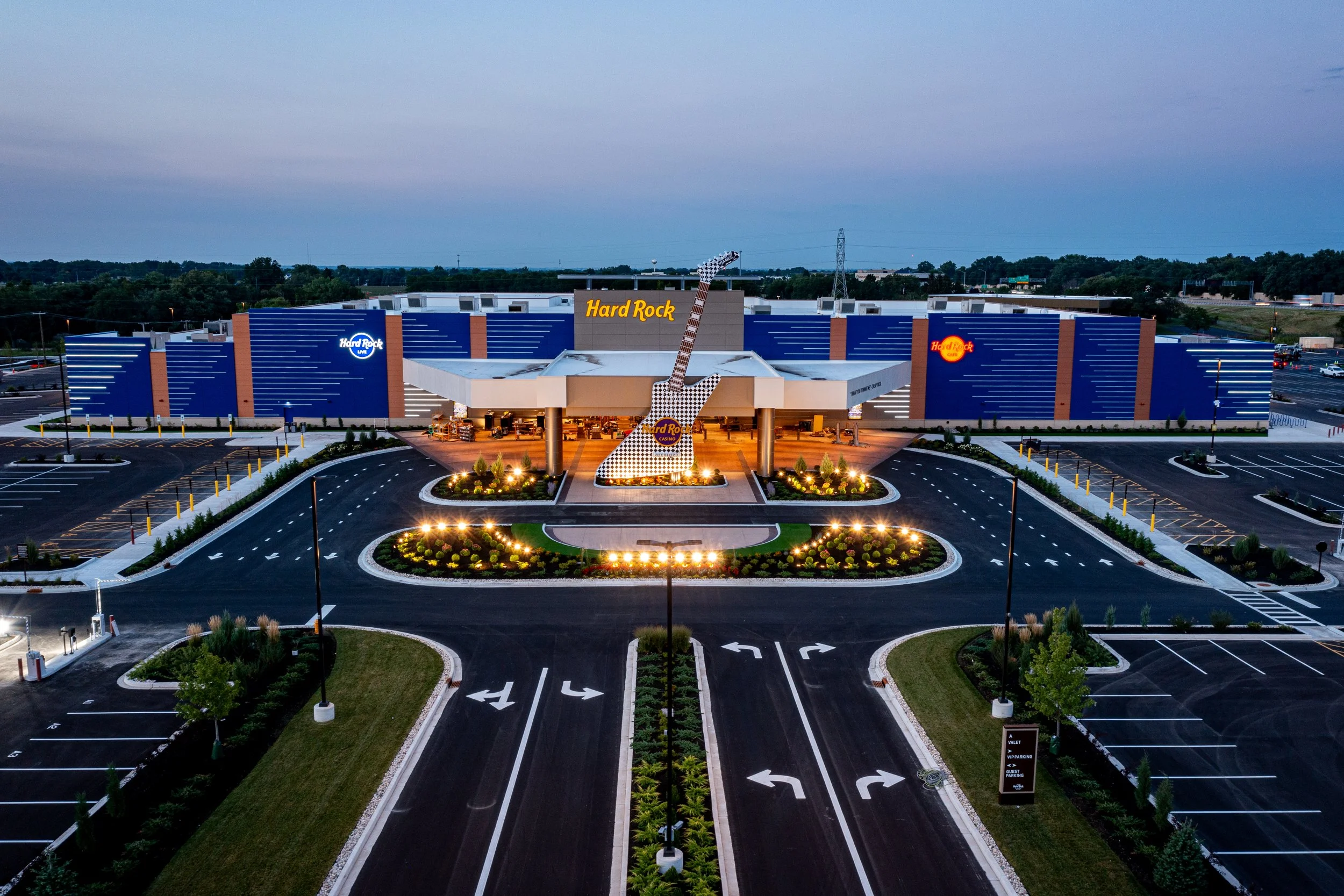 Exterior view of the Hard Rock casino with a large guitar sculpture in front, illuminated at dusk, surrounded by a parking lot with marked spaces and landscaped areas.