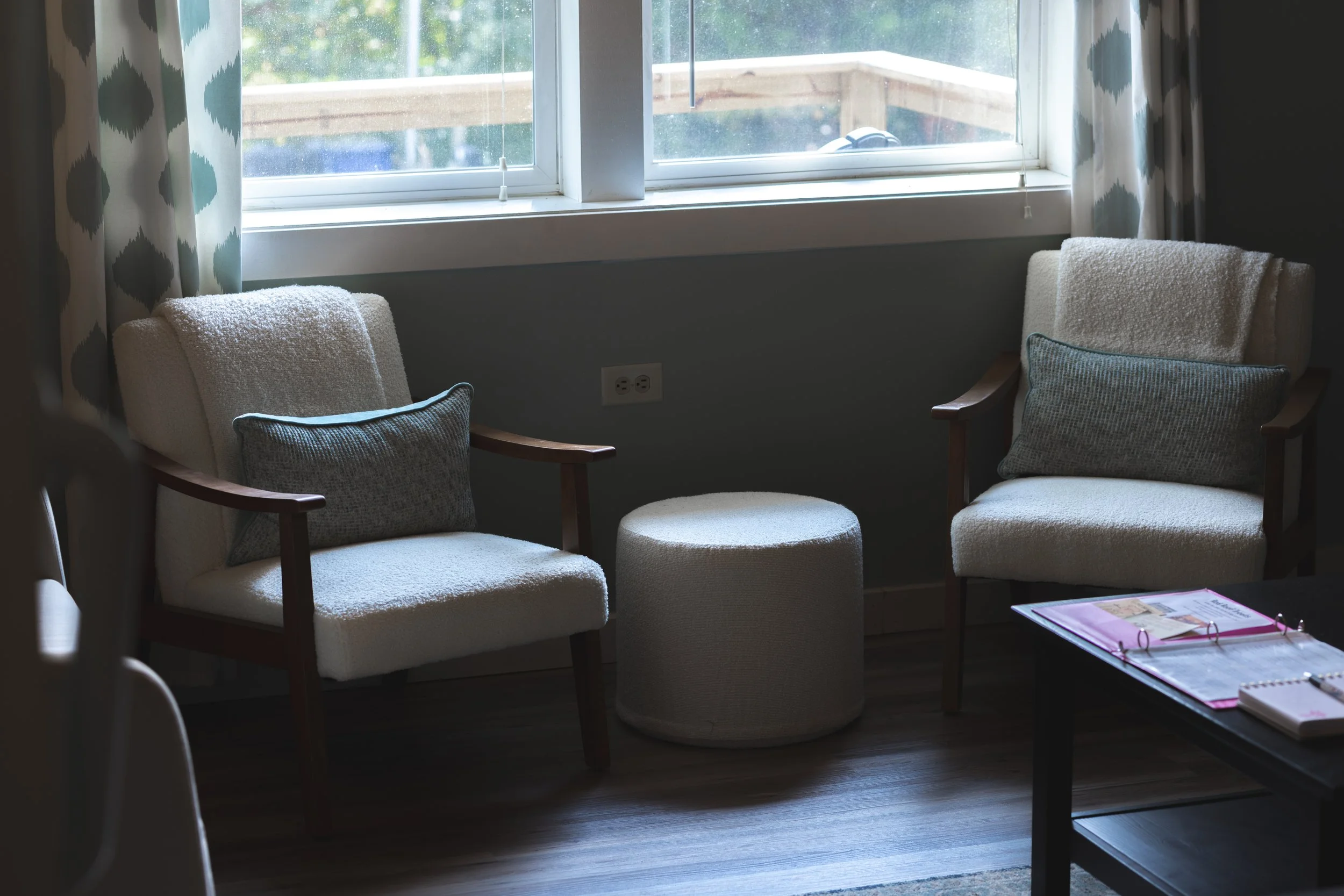 Two beige armchairs with wooden arms and gray pillows are positioned near a window in a living room. A small round beige ottoman is between the chairs. A black coffee table with pink binders and notebooks is in the foreground. Blue and white patterned curtains hang beside the window.