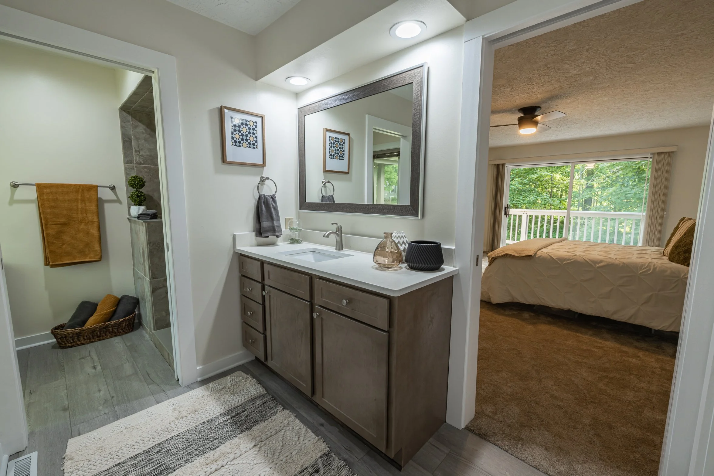 Bathroom with vanity mirror, sink, and cabinets, adjacent to a bedroom with large window and bed, and a separate shower area with towels and decorative items.