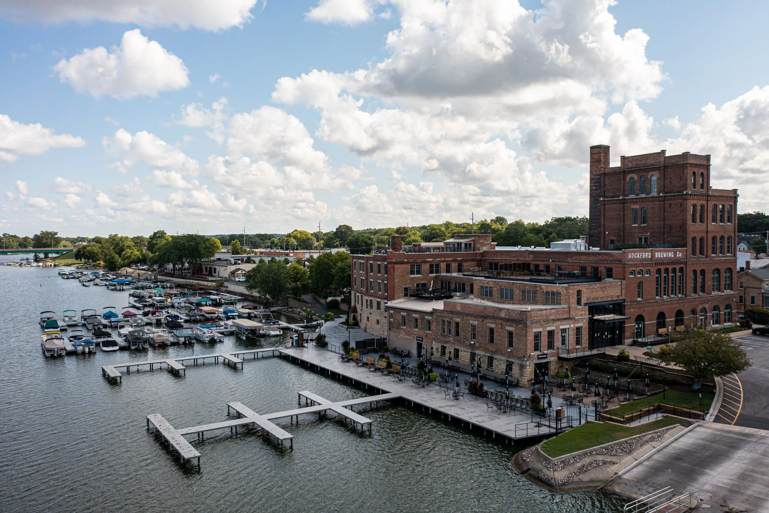 A waterfront scene featuring a brick building labeled 'Rockford Brewing Co,' a dock with boats, and a parking lot with cars. Trees line the background, and the sky is blue with white clouds.