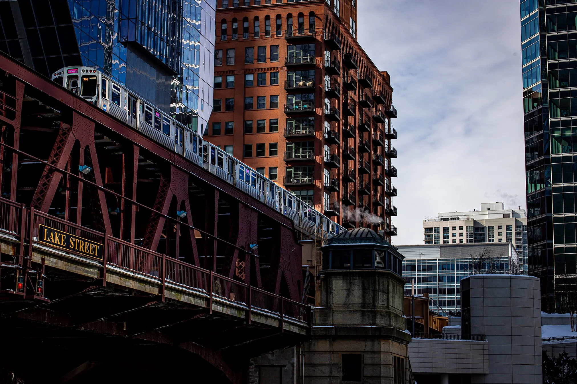 An elevated train on a red steel bridge passing through a city with tall modern buildings, and a sign reading 'Lake Street' on the bridge.
