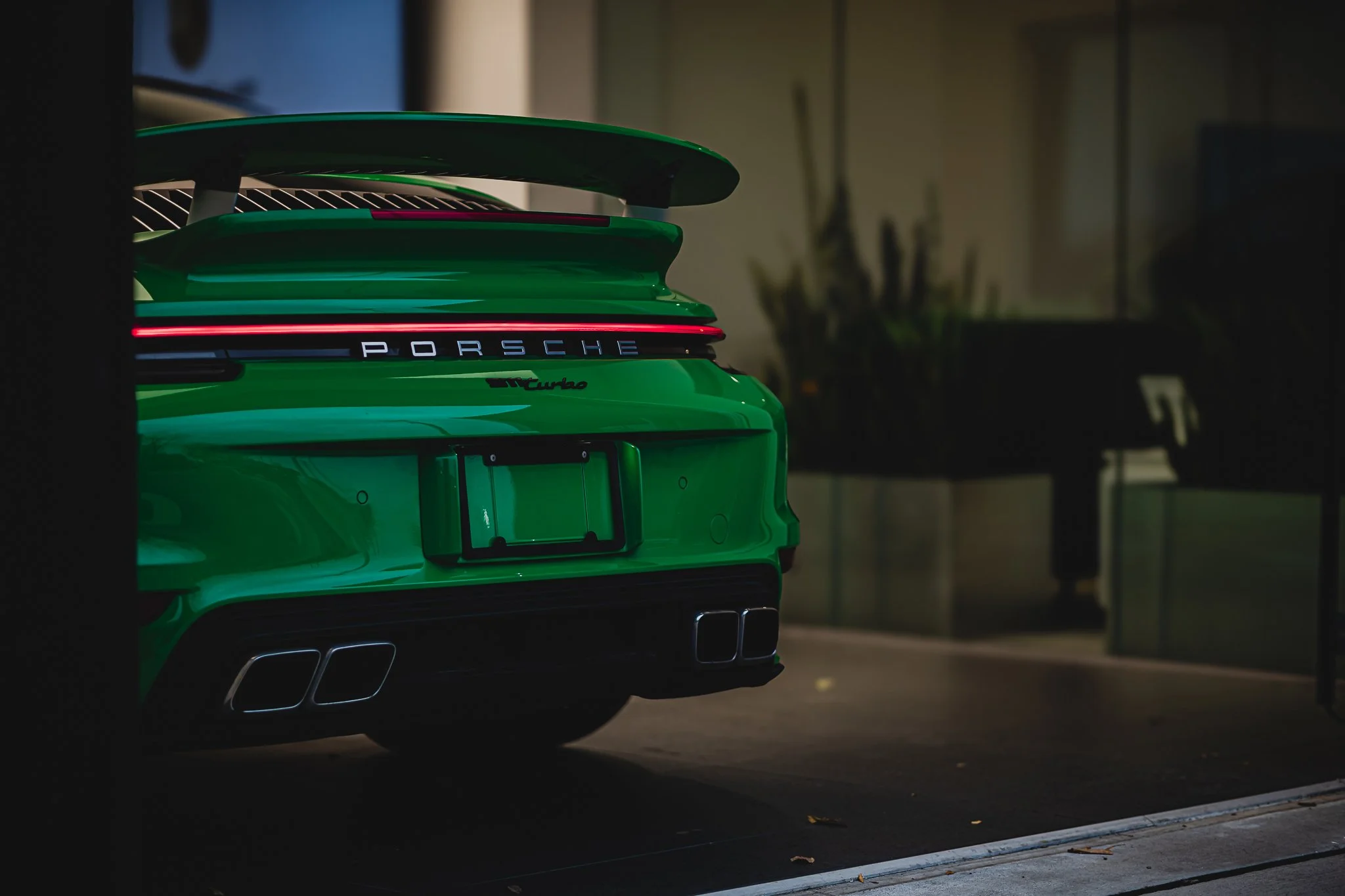 The rear of a bright green Porsche 911 Turbo car inside a garage, showing the Porsche logo, model name, dual exhaust pipes, and a red taillight strip.