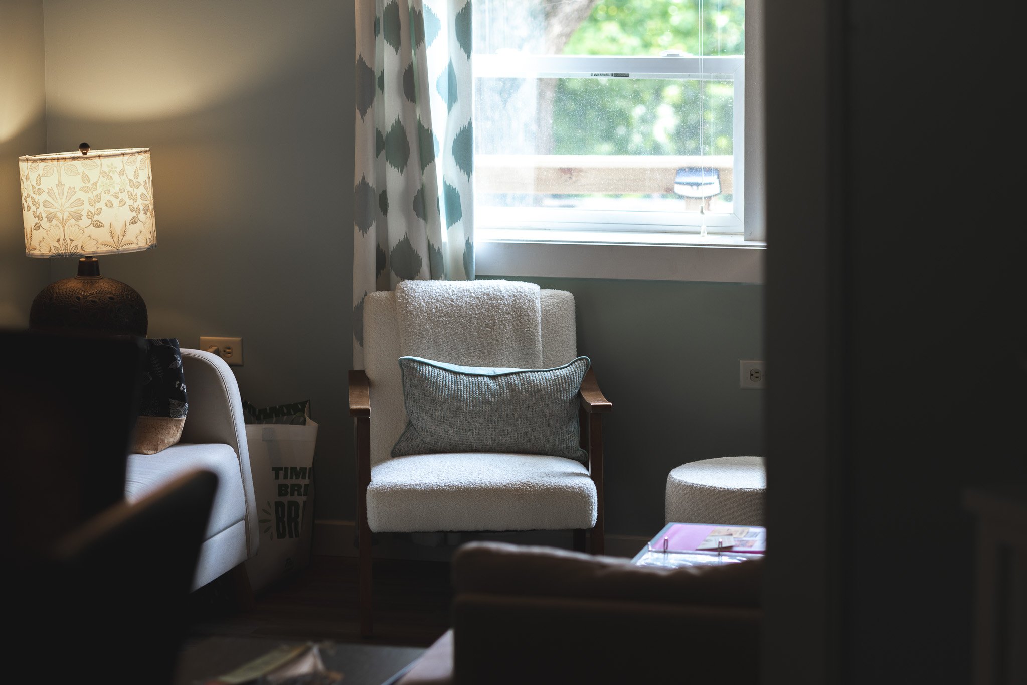 A cozy living room corner with a white armchair, pillow, and a window with patterned curtains, a table lamp, and a small table.