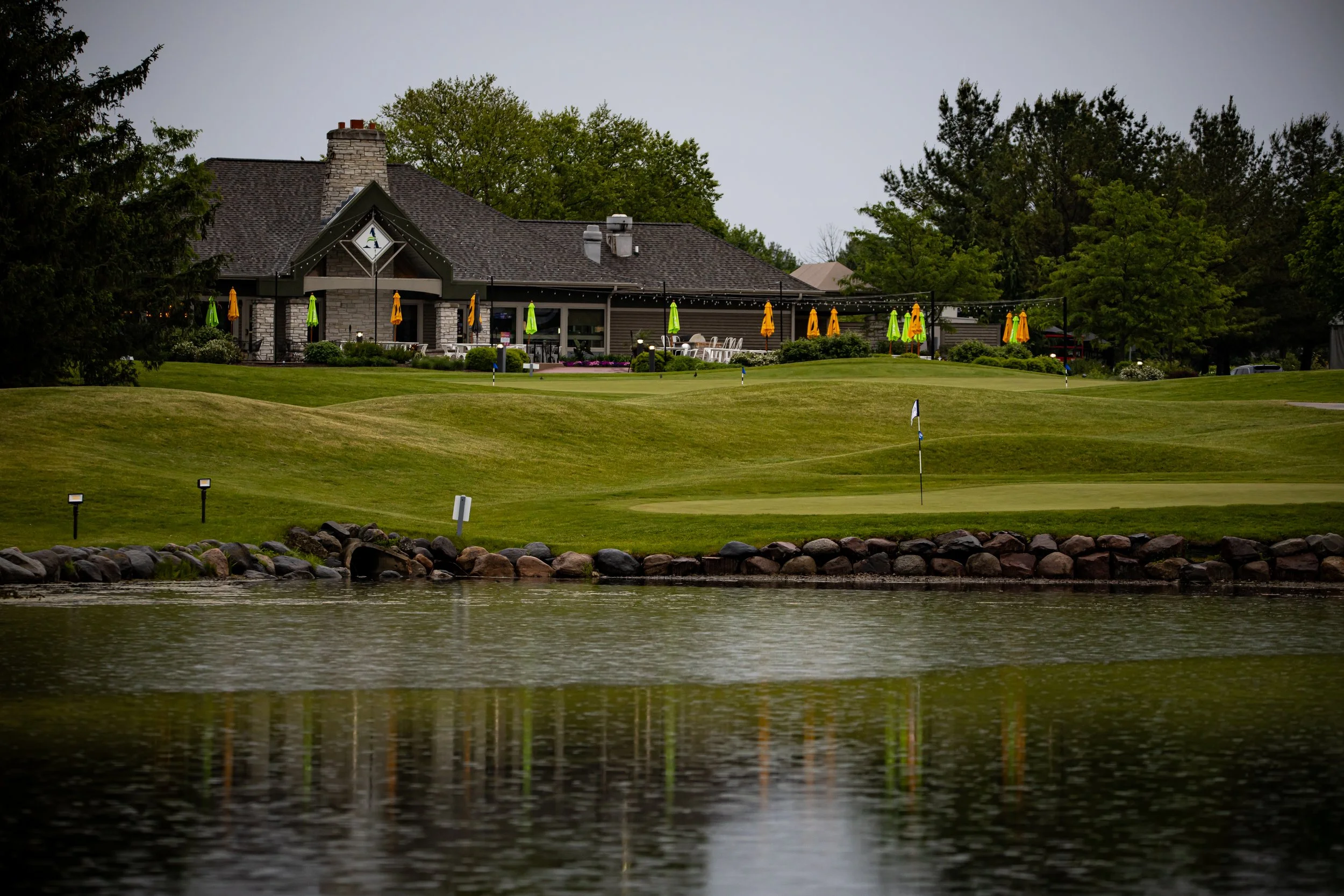 A golf course with a putting green and flag, surrounded by rocks and a body of water, with a clubhouse in the background, decorated with yellow and green umbrellas and trees under an overcast sky.